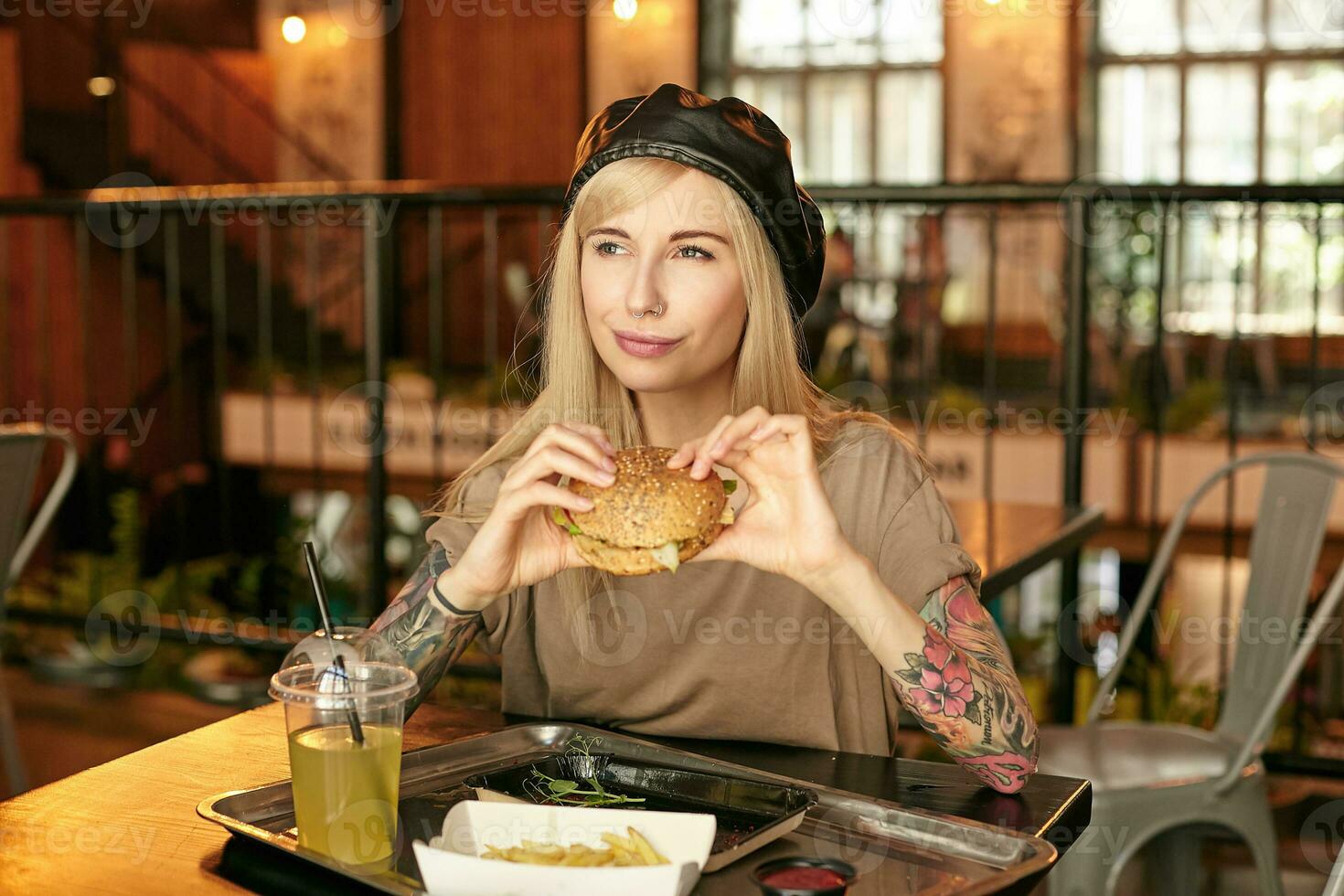 Young satisfied blonde long haired lady with tattooes posing over modern cafe interior and holding hamburger in hands, looking aside and smiling happily, wearing trendy clothes photo