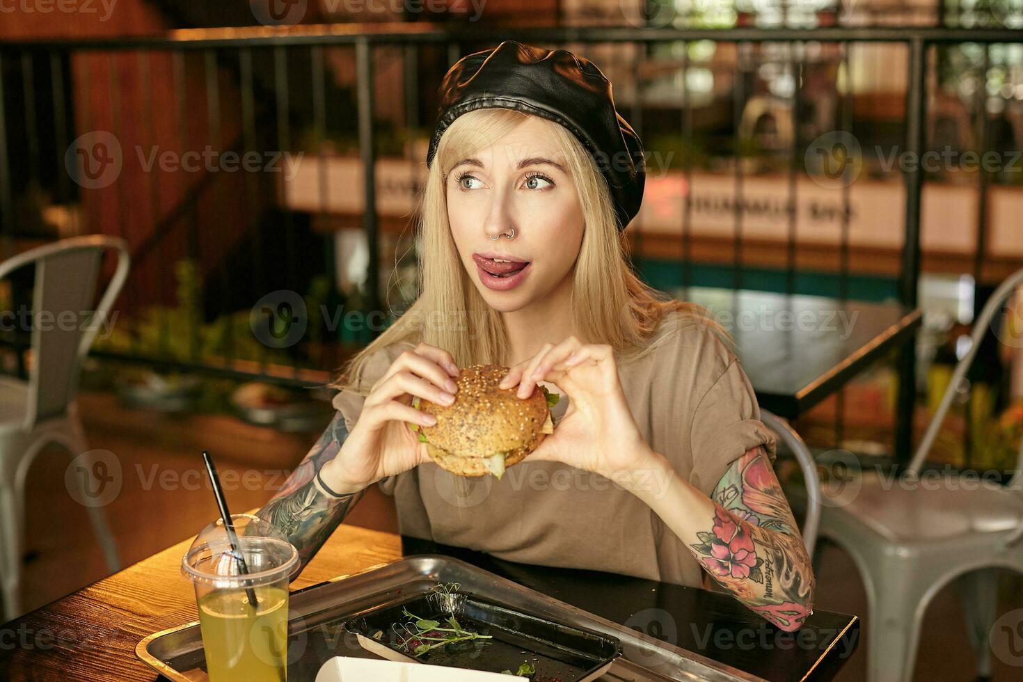 Portrait of attractive young tattooed blonde woman in beige t-shirt and black beret eating hamburger and licking her lips, posing over modern cafe interior photo