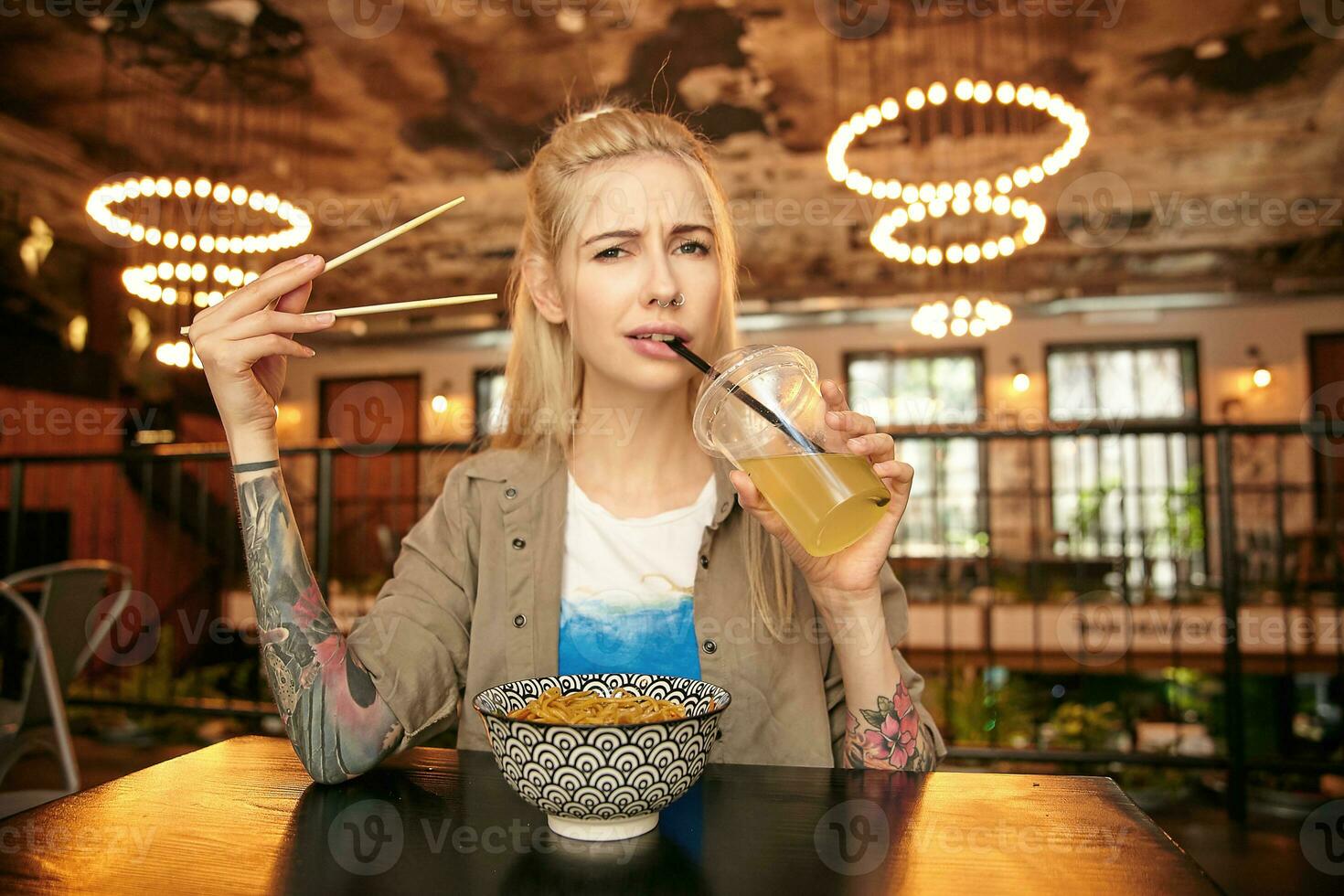 Portrait of pretty young long haired female with tattooes sitting at table in modern cafe, holding wooden sticks in hand and drinking lemonade with straw, wearing casual clothes photo