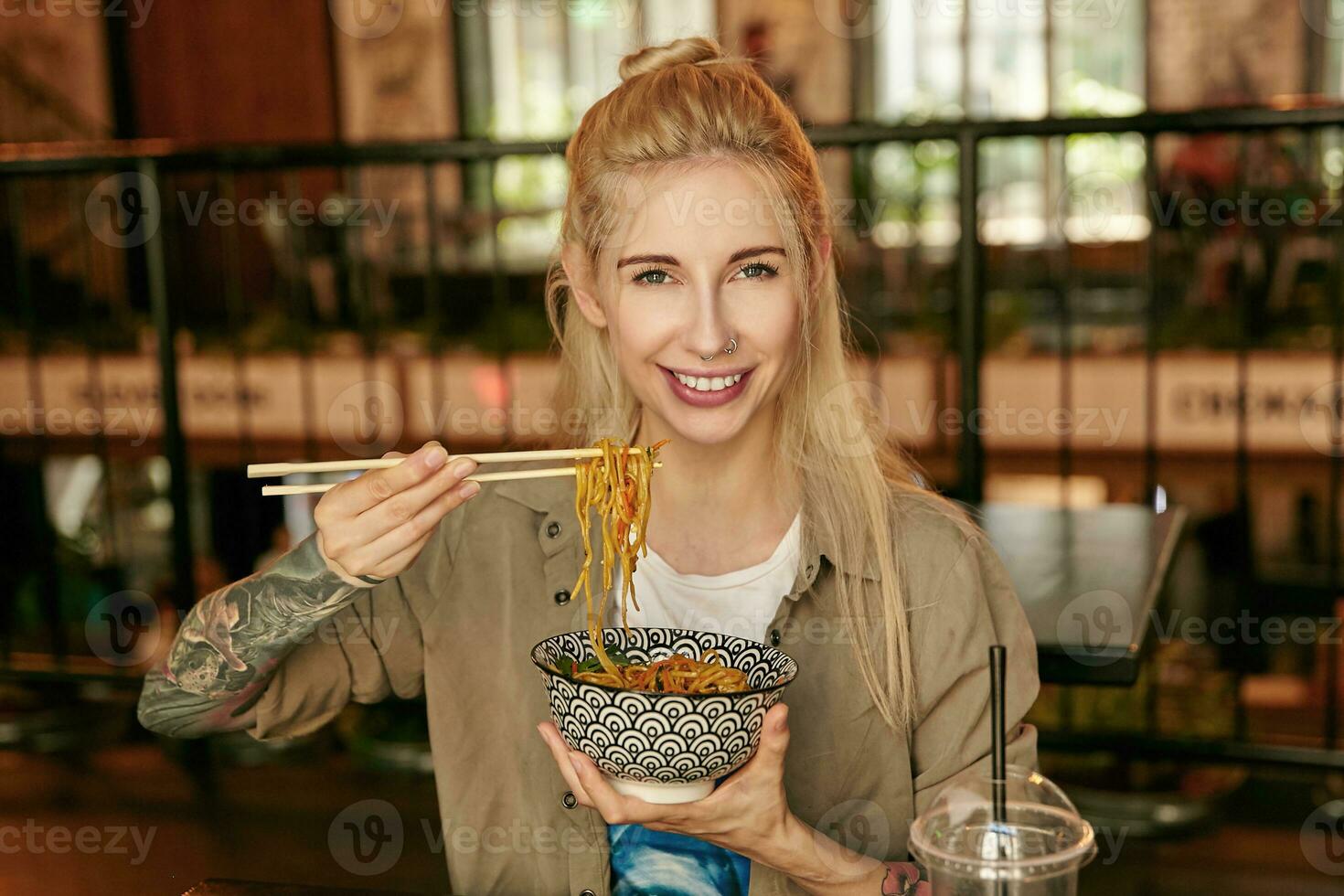 Cheerful young tattooed blonde woman in casual clothes posing over restaurant interior, holding bowl with noodles and looking at camera with wide happy smile photo