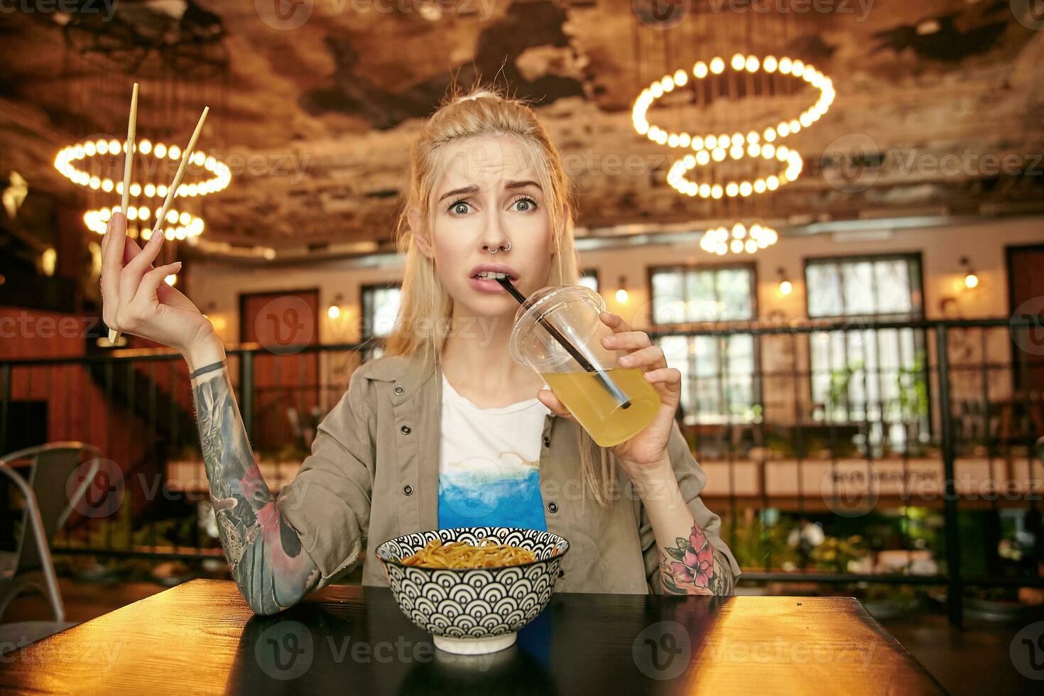 Puzzled young tattooed blonde female posing over city cafe interior in casual clothes, sitting at table with asian food and drinking lemonade, rounding eyes and frowning while looking to camera photo