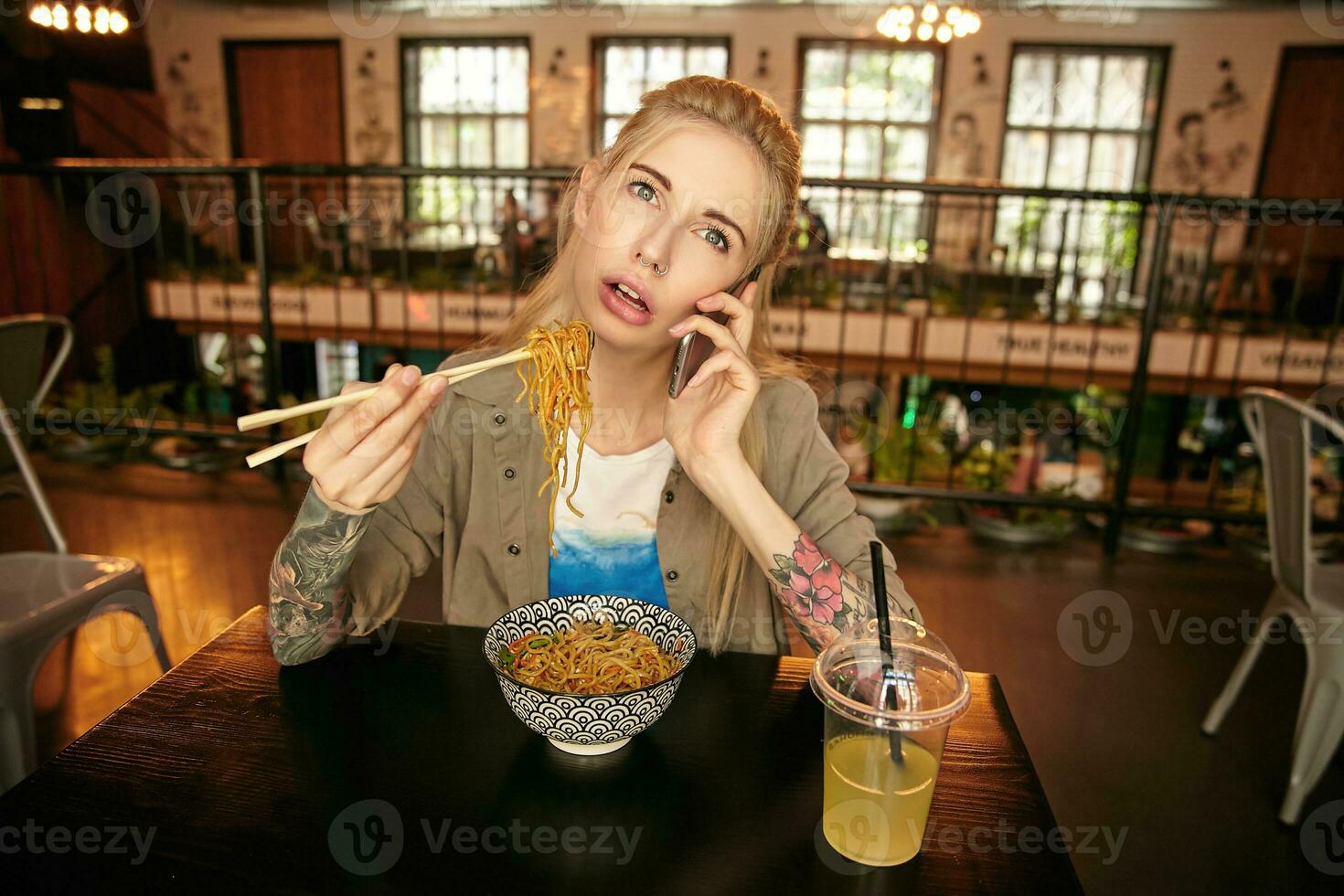 Horizontal shot of pretty young blonde woman with tattooes having linch in modern cafe, wearing beige shirt and white t-shirt, having boring conversation on her phone photo