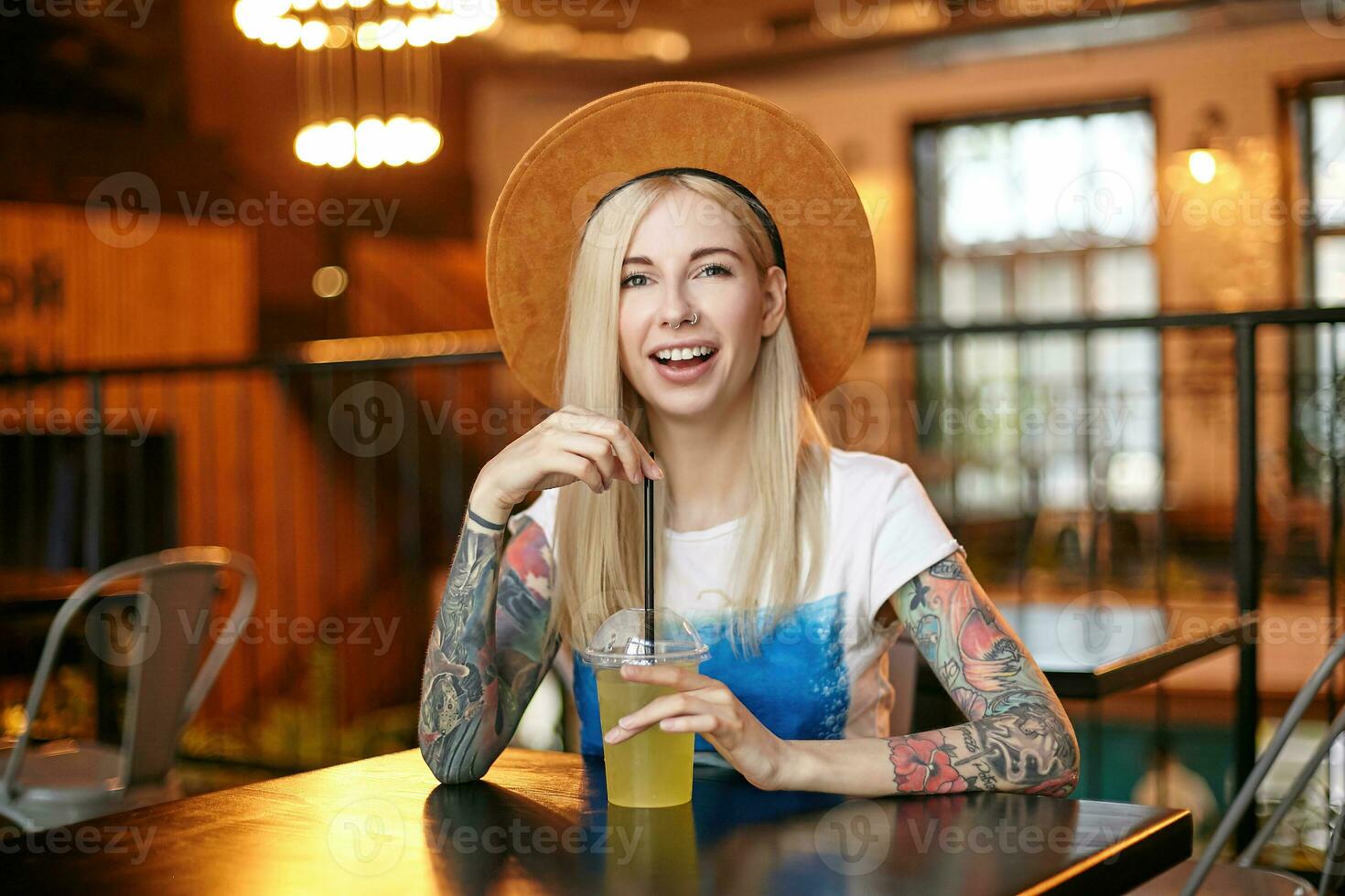 Cheerful blonde long haired woman with tattooes keeping hands on table while drinking fresh juice with straw, looking at camera positively and smiling joyfully photo