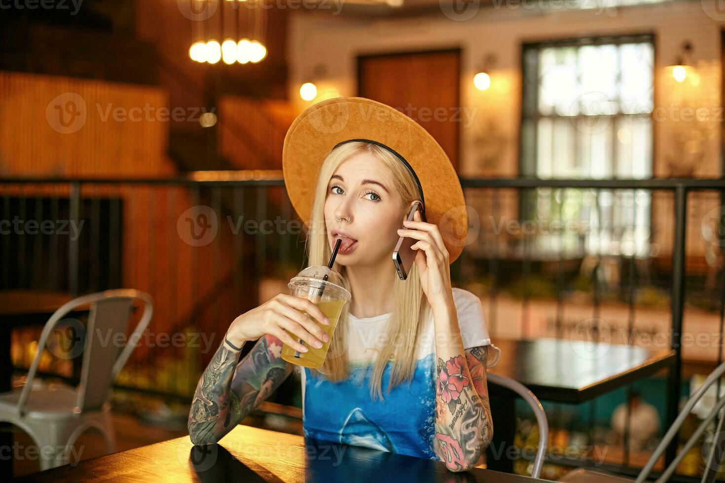 Indoor photo of pretty young blonde long haired female making call with her smartphone and looking at camera cheerfully, sitting at table in city cafe and drinking lemonade