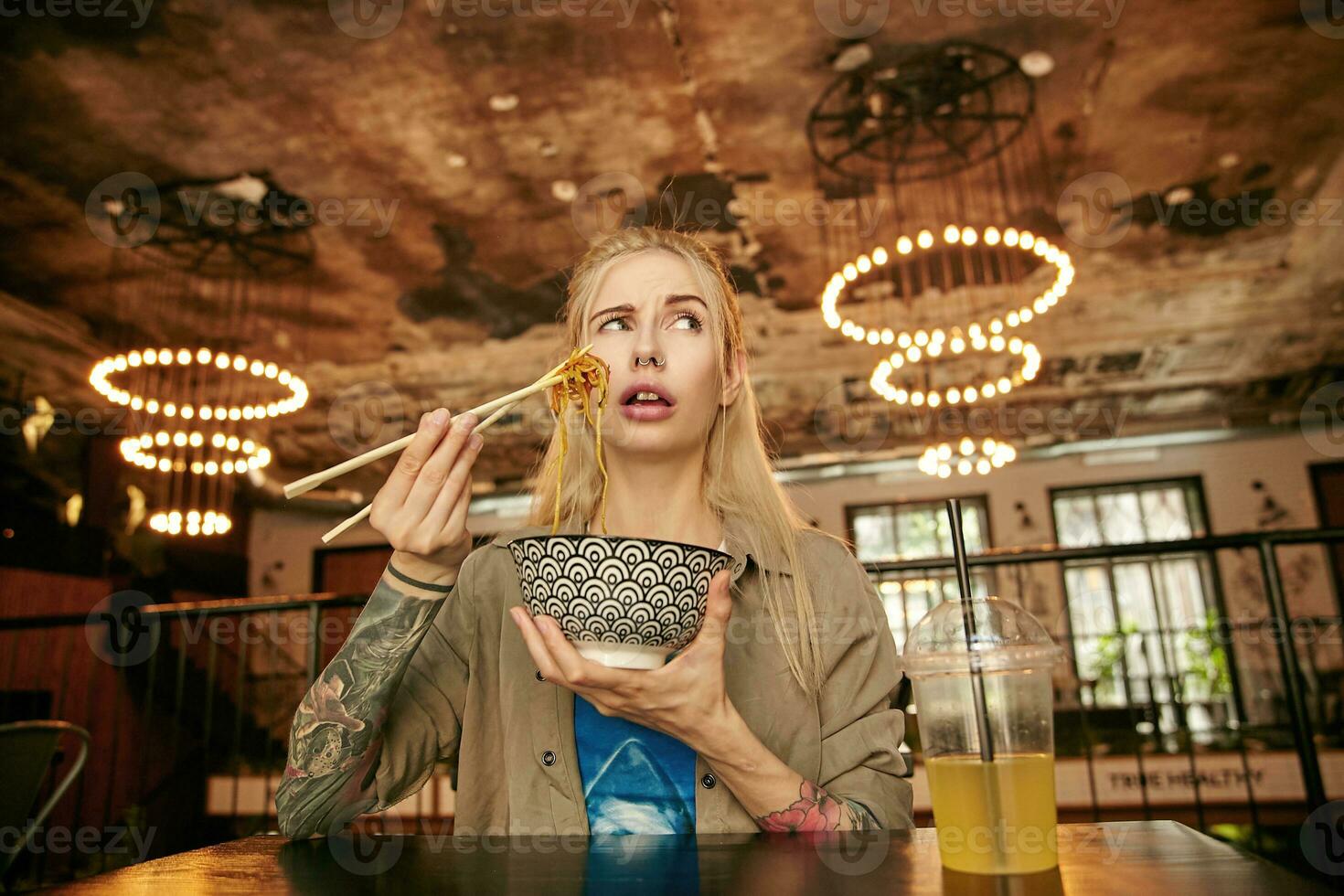 Portrait of doubting tattooed blonde female with casual hairstyle trying first asian food, holding caramic bowl with noodles and looking aside with confused face photo