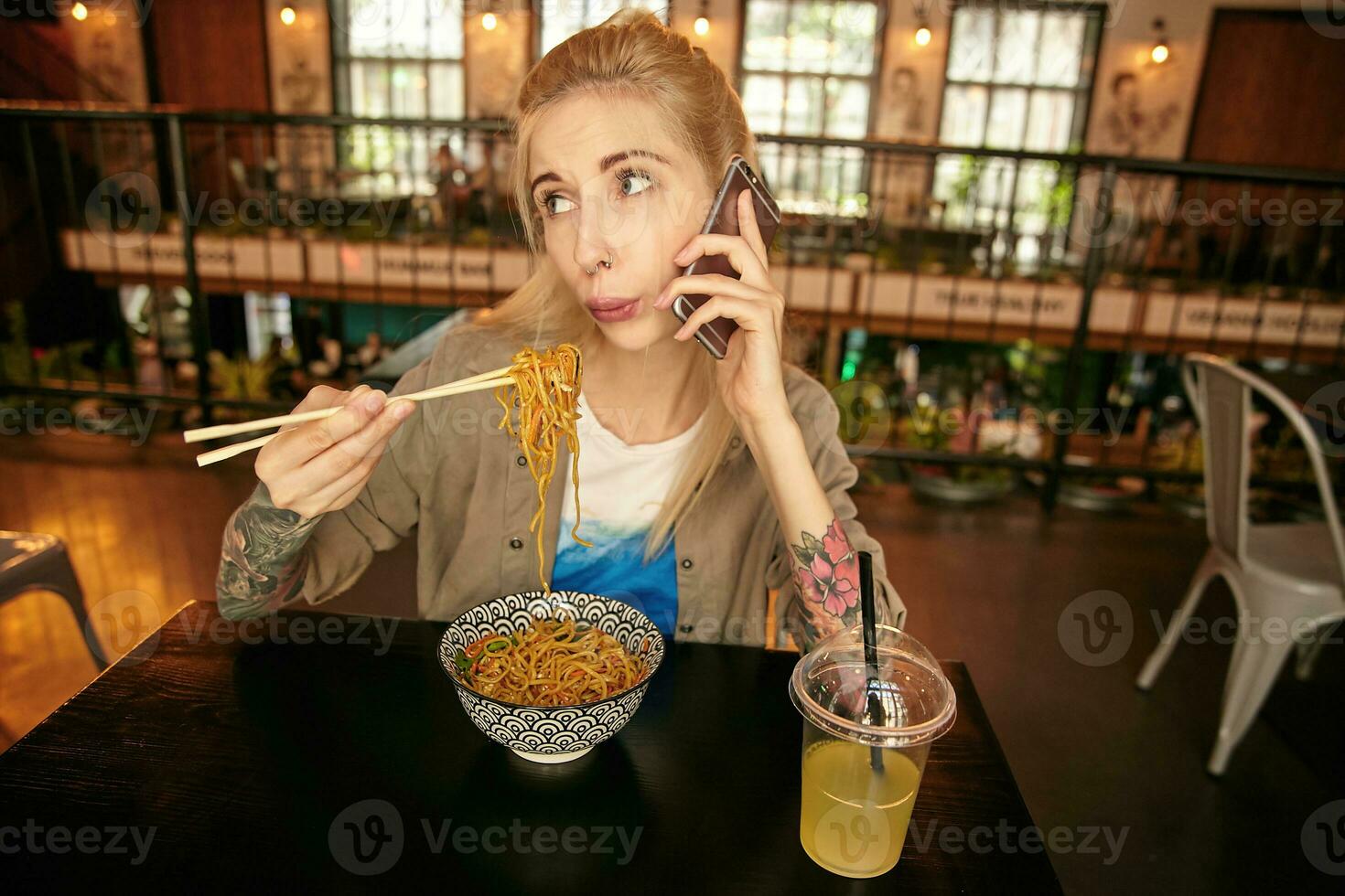 Indoor photo of pretty tattooed blonde woman with casual hairstyle eating asian food with wooden sticks, blowing on hot food while making call with her mobile phone