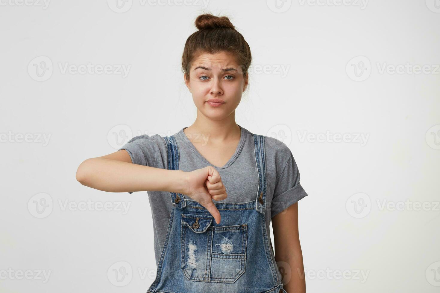 Unimpressed disliking attractive female student in denim overall, showing thumb down and smirking from displeasure, giving negative feedback about concert, frowning and standing over white wall photo