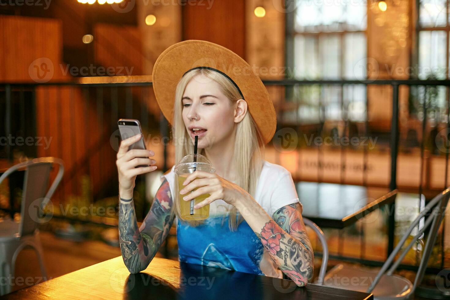 Portrait of pretty young long haired female with tattooes sitting at table in cafe with modern interior and drinking lemonade, holding smartphone in raised hand and looking at screen photo