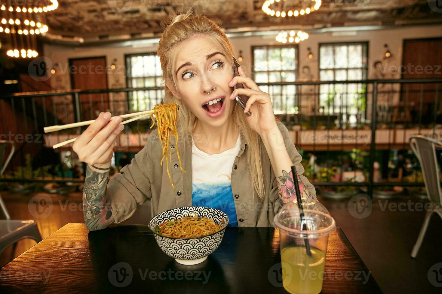 Young blonde female with tattooes having excited talk on her phone while eating noodles with wooden sticks in modern restaurant, wrinkling forehead and looking aside with wide mouth opened photo