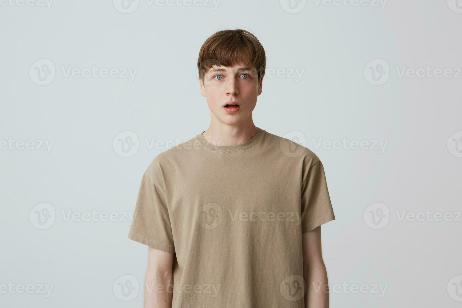 Portrait of amazed handsome young man in beige t shirt with short haircut and opened mouth standing and looks surprised isolated over white background photo