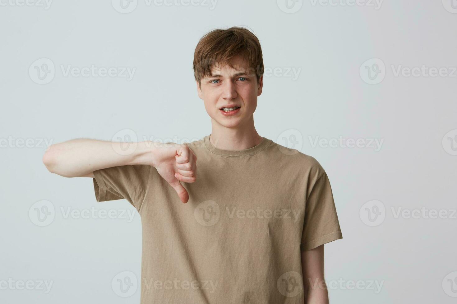 Closeup of sad disappointed young man in beige t shirt with braces on teeth looks upset and showing thumbs down isolated over white background photo