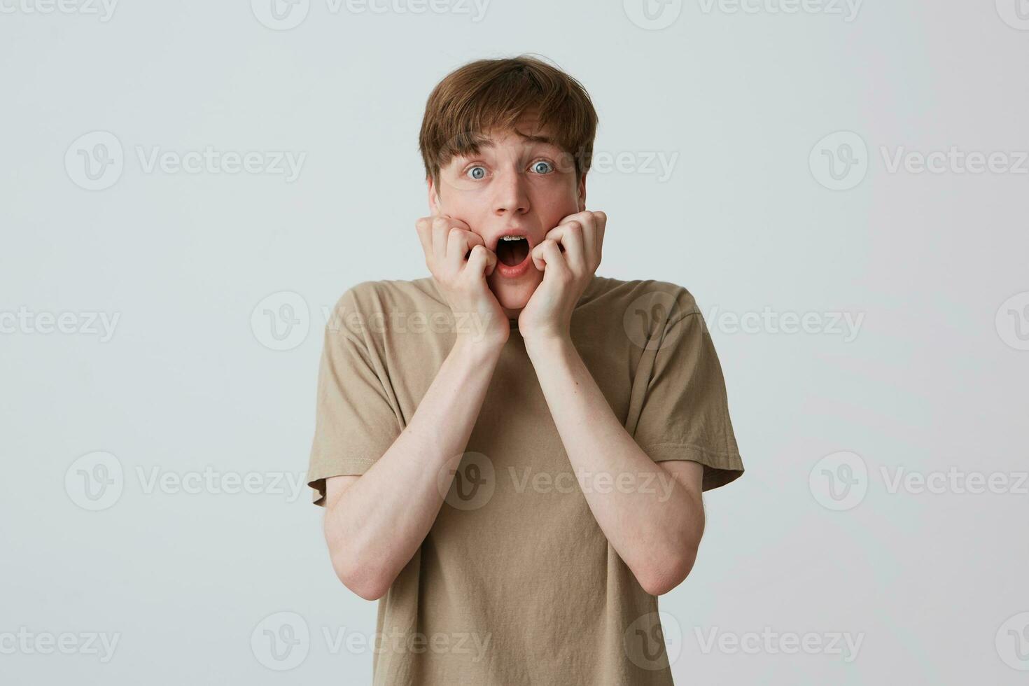 Closeup of scared shocked young man in beige t shirt with short haircut and opened mouth looks frightened and shouting isolated over white background photo