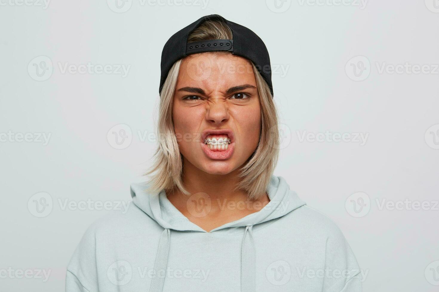 Portrait of mad angry young woman with braces wears black cap and blue hoodie looks irritated and shows teeth isolated over white background photo