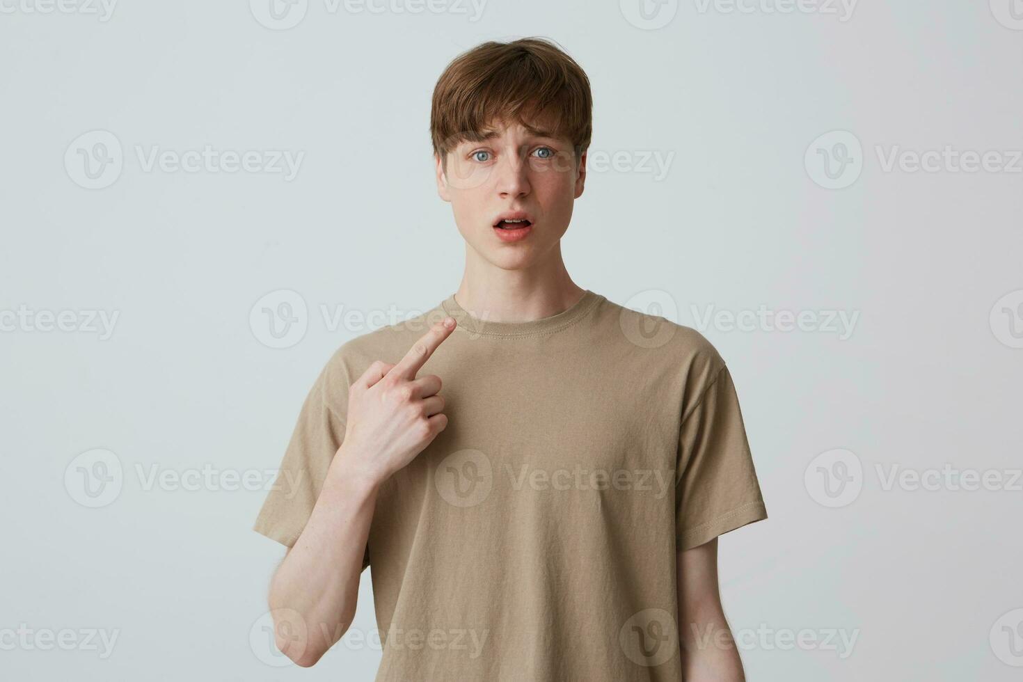 Portrait of sad amazed young man with short haircut in beige t shirt looks shocked and pointing at himself with finger isolated over white background photo