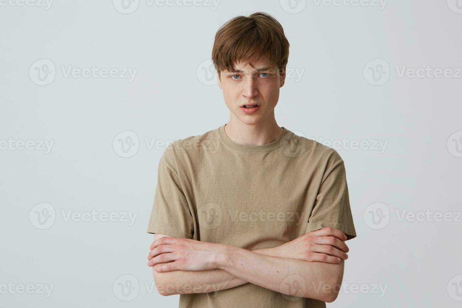 retrato de trastornado infeliz joven hombre con corto Corte de pelo en beige t camisa mira desesperado y en pie con brazos cruzado aislado terminado blanco antecedentes foto