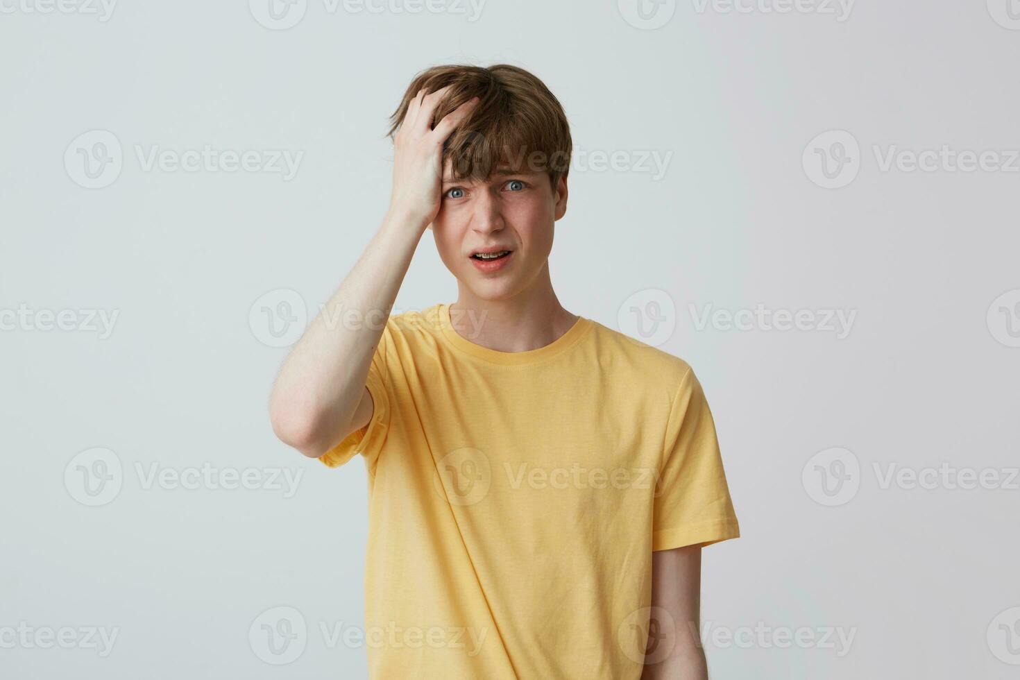 retrato de desesperado infeliz joven hombre con mano en cabeza y tirantes en dientes en amarillo t camisa mira triste y teniendo un dolor de cabeza aislado terminado blanco antecedentes foto