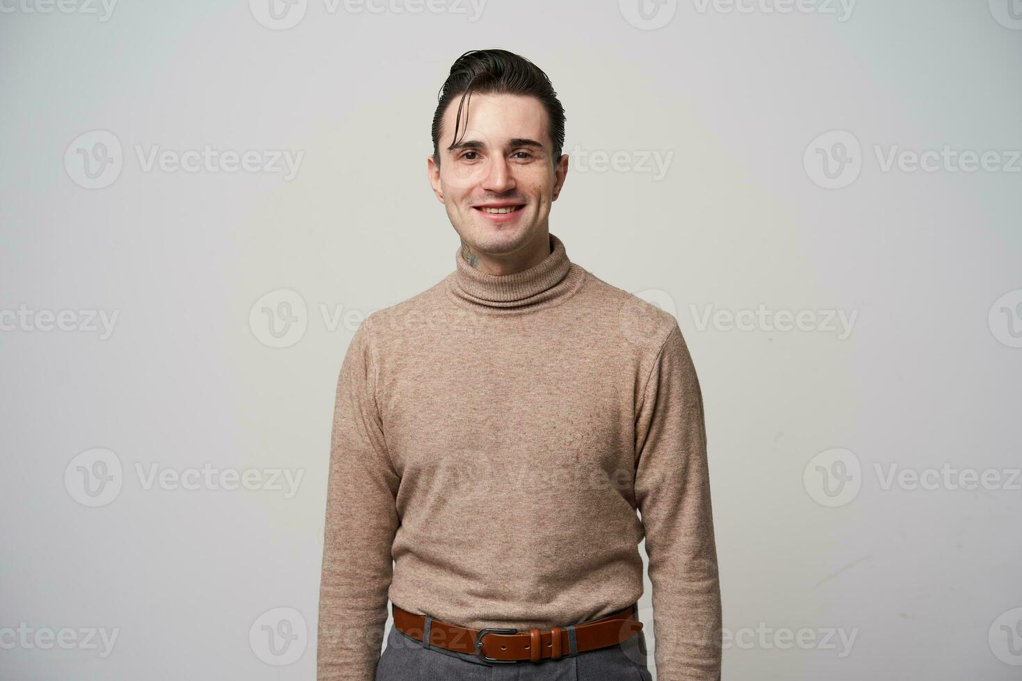 Positive pretty young dark haired male with trendy haircut looking at camera and smiling slightly, standing over white background with hands down, dressed in fashionable clothes photo