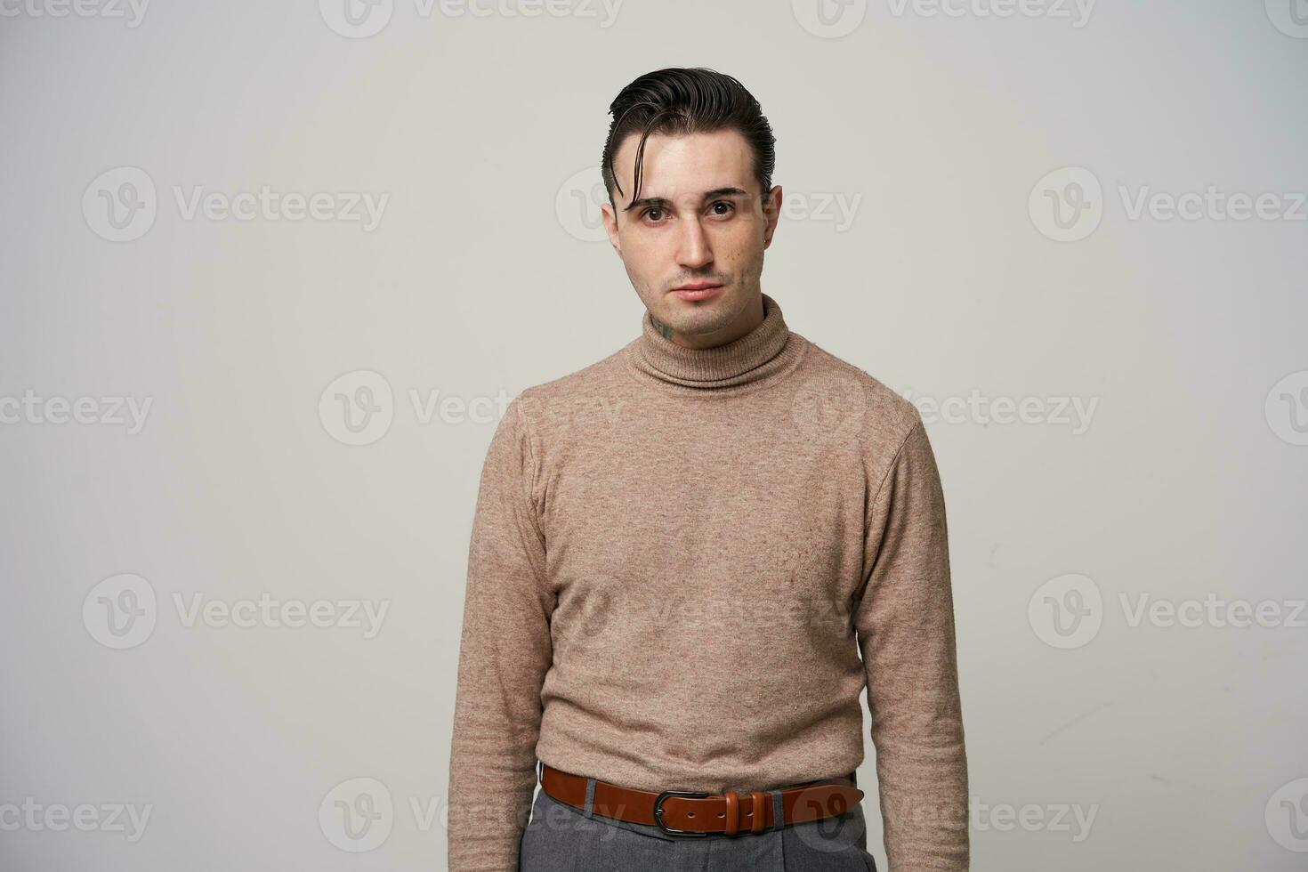 Serious brow-eyed handsome young brunette male with trendy haircut wearing beige roll-neck sweater and grey trousers while posing over white background photo