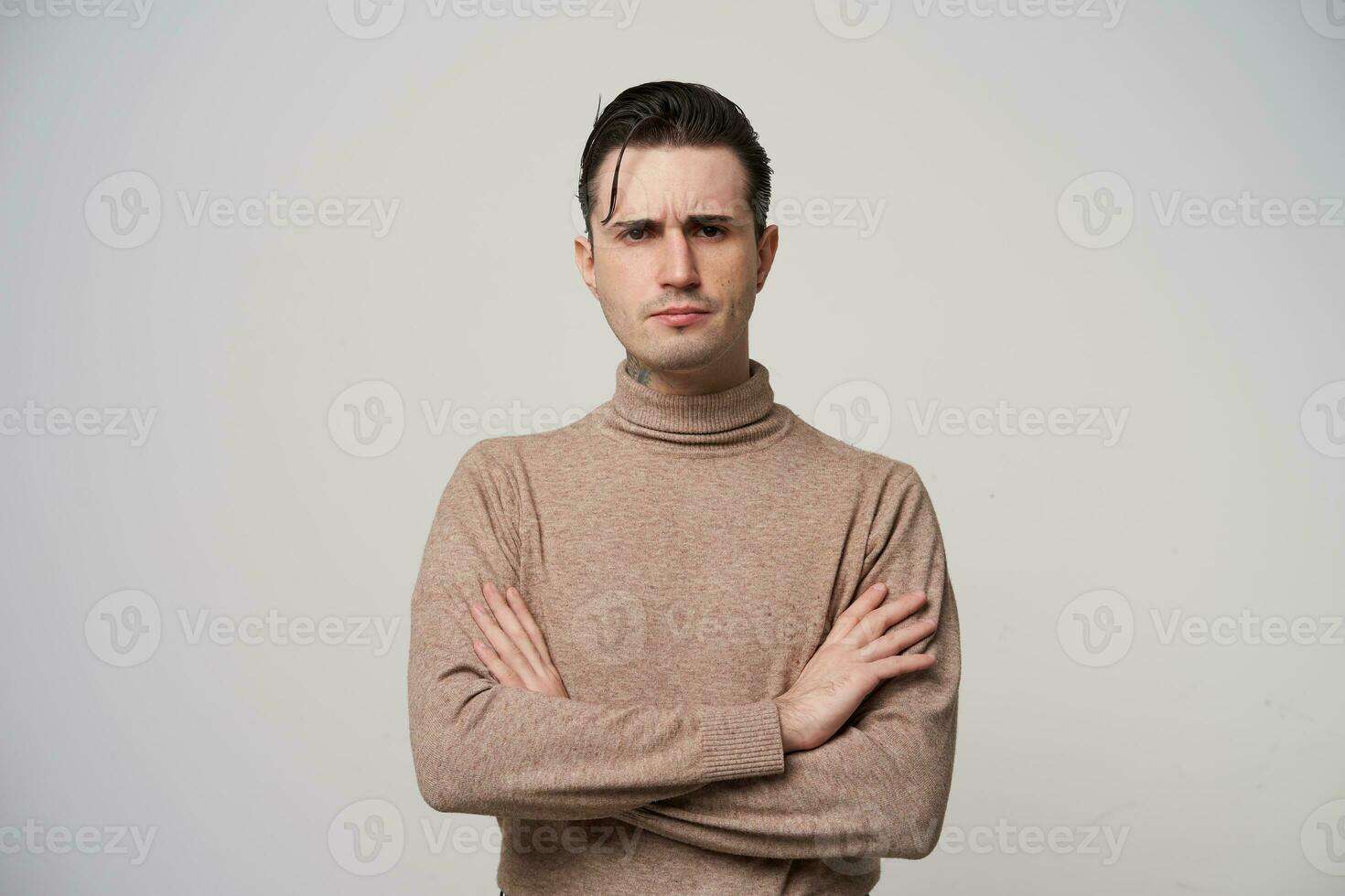 retrato de hermoso joven morena masculino en de moda vestir vistiendo elegante peinado, plegable manos en su cofre mientras posando terminado blanco fondo, mirando seriamente a cámara y ceñudo Cejas foto