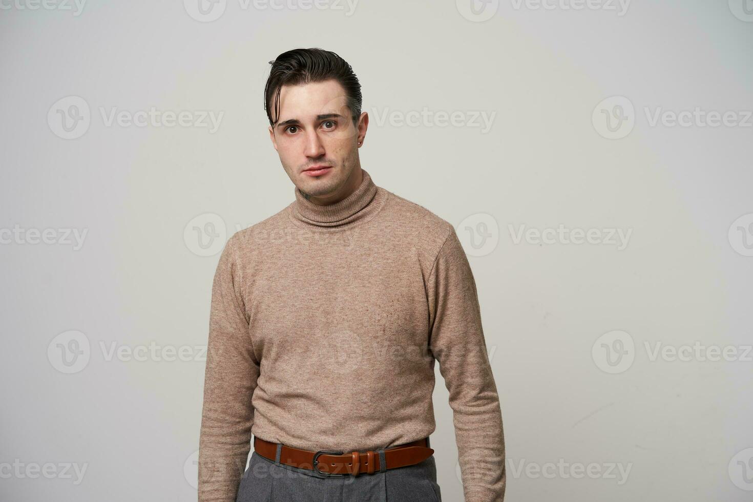 estudio foto de atractivo joven oscuro peludo masculino hipster con elegante peinado mirando y cámara con doblada labios, participación manos a lo largo su cuerpo mientras en pie terminado blanco antecedentes