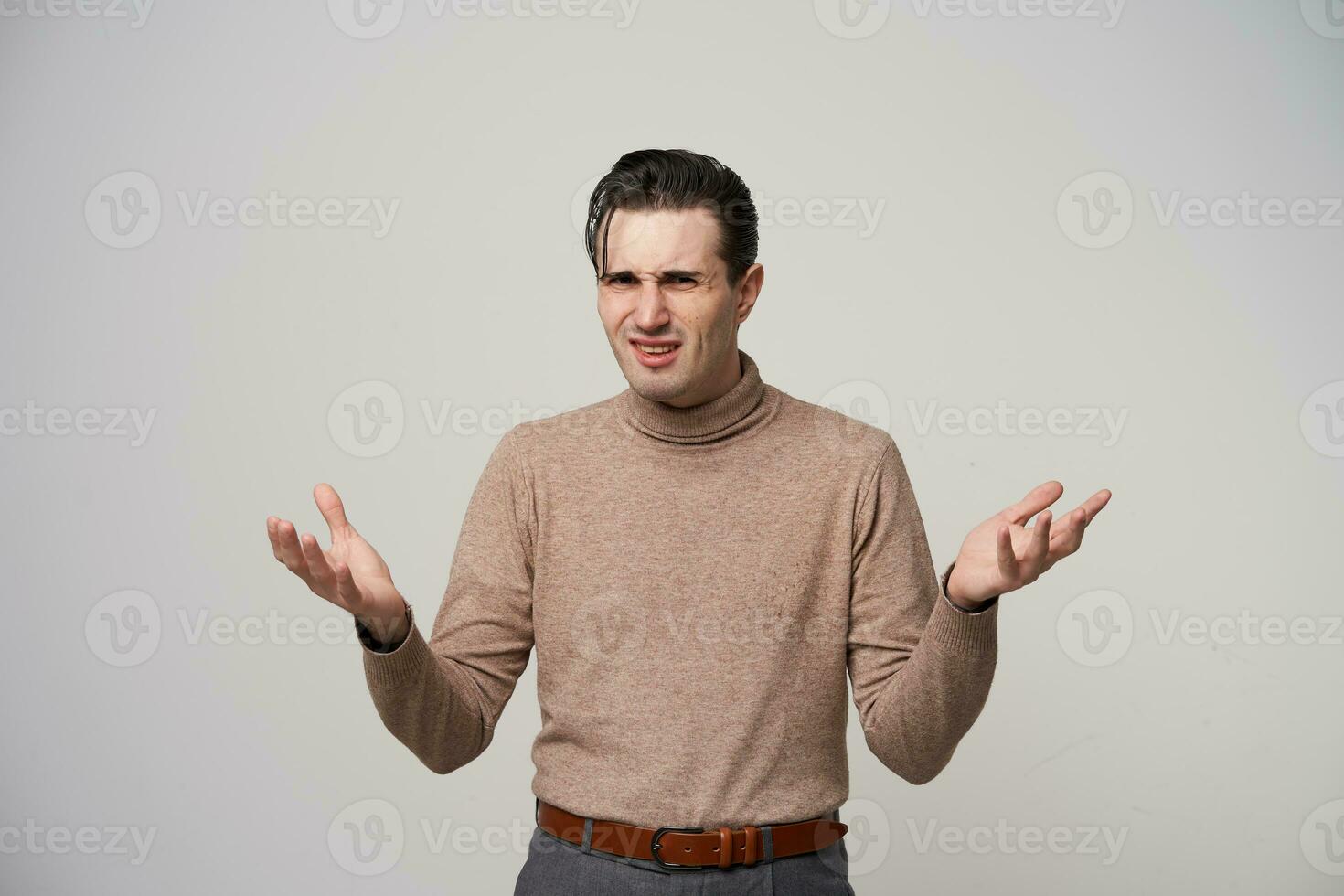 Puzzled young dark haired man in beige roll-neck sweater looking to camera with pout and frowning his face with raised palms, standing against white background photo