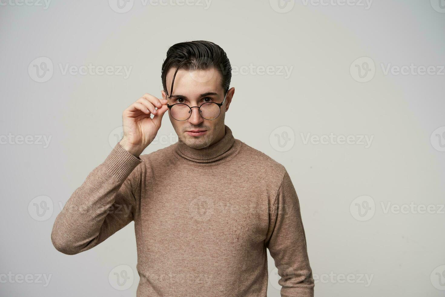 joven de ojos marrones hermoso morena chico participación mano en su gafas y mirando atentamente a cámara con doblada labios, vestido en de moda vestir mientras posando terminado blanco antecedentes foto