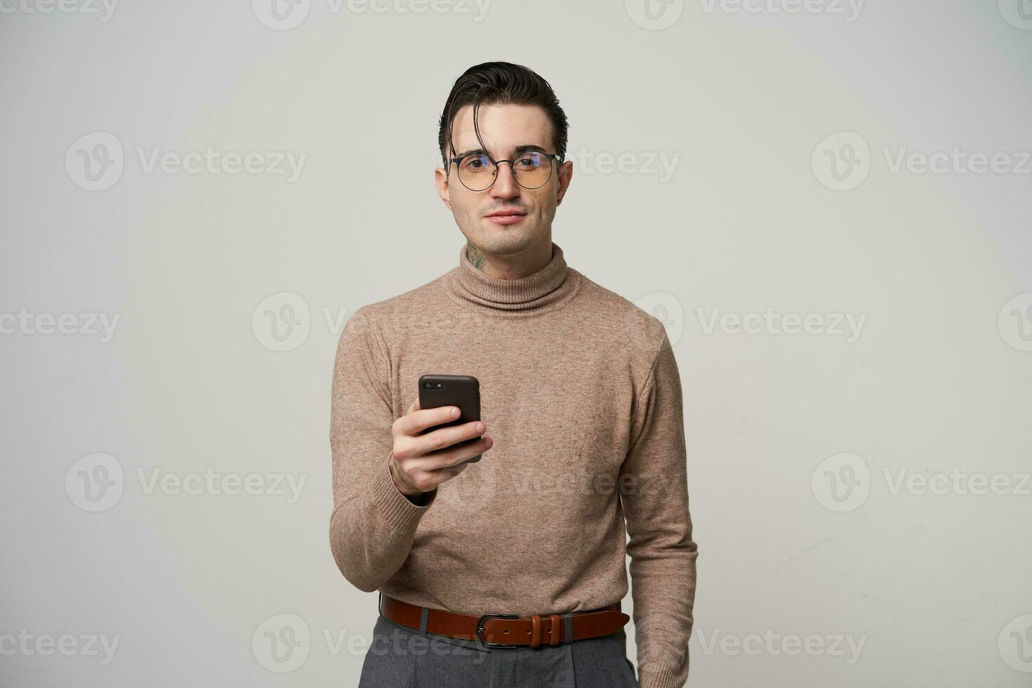 Positive young pretty brunette male hipster with stylish hairstyle holding smartphone and looking at camera with light smile, standing over white background in glasses photo