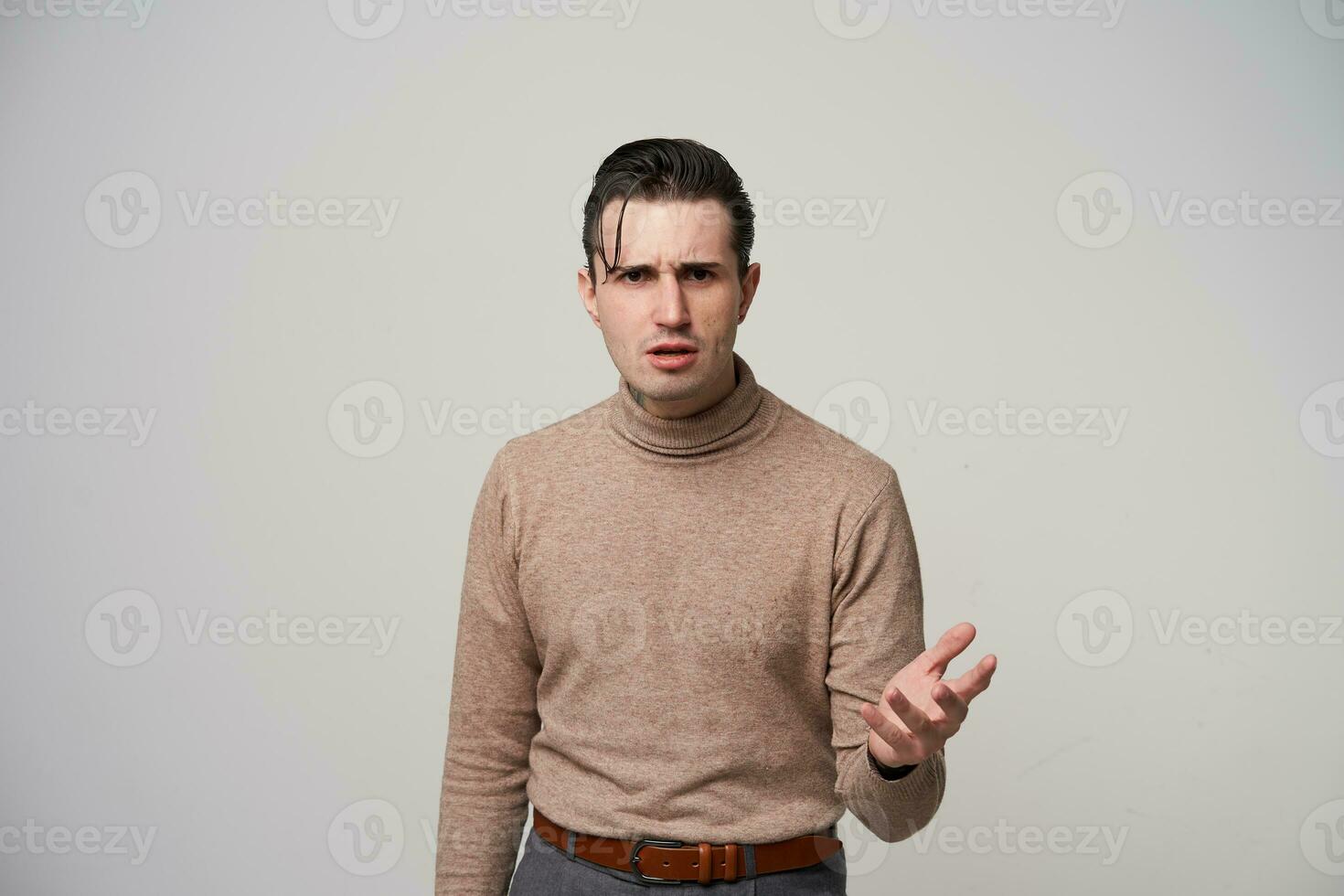 Perplexed young dark haired male hipster with stylish hairstyle looking confusedly to camera and frowning his eyebrows, posing over white background with raised hand photo