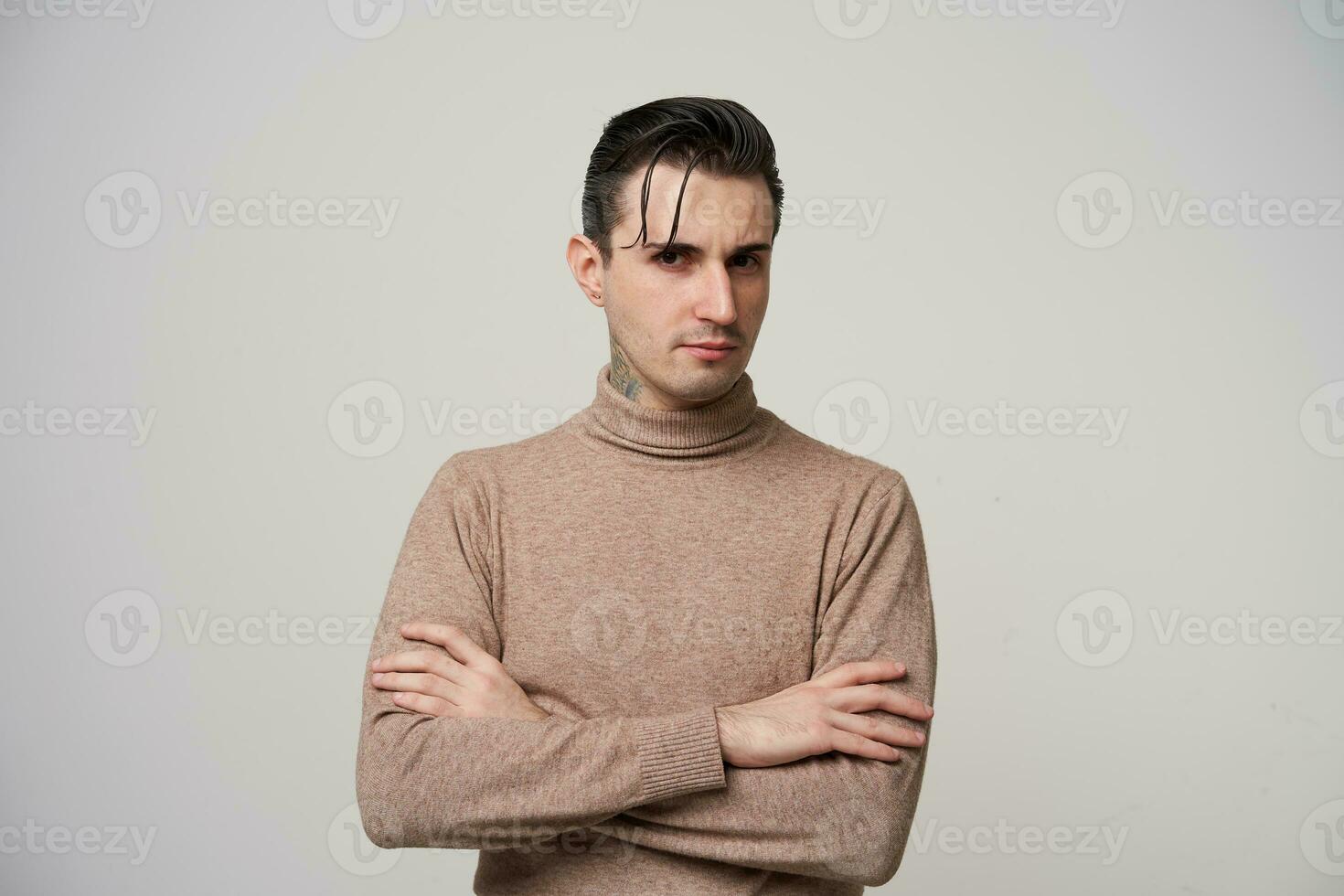 Studio shot of severe young brunette male hipster with tattoo on his neck standing over white background in fashionable clothes, looking seriously to camera and frowning eyebrows photo