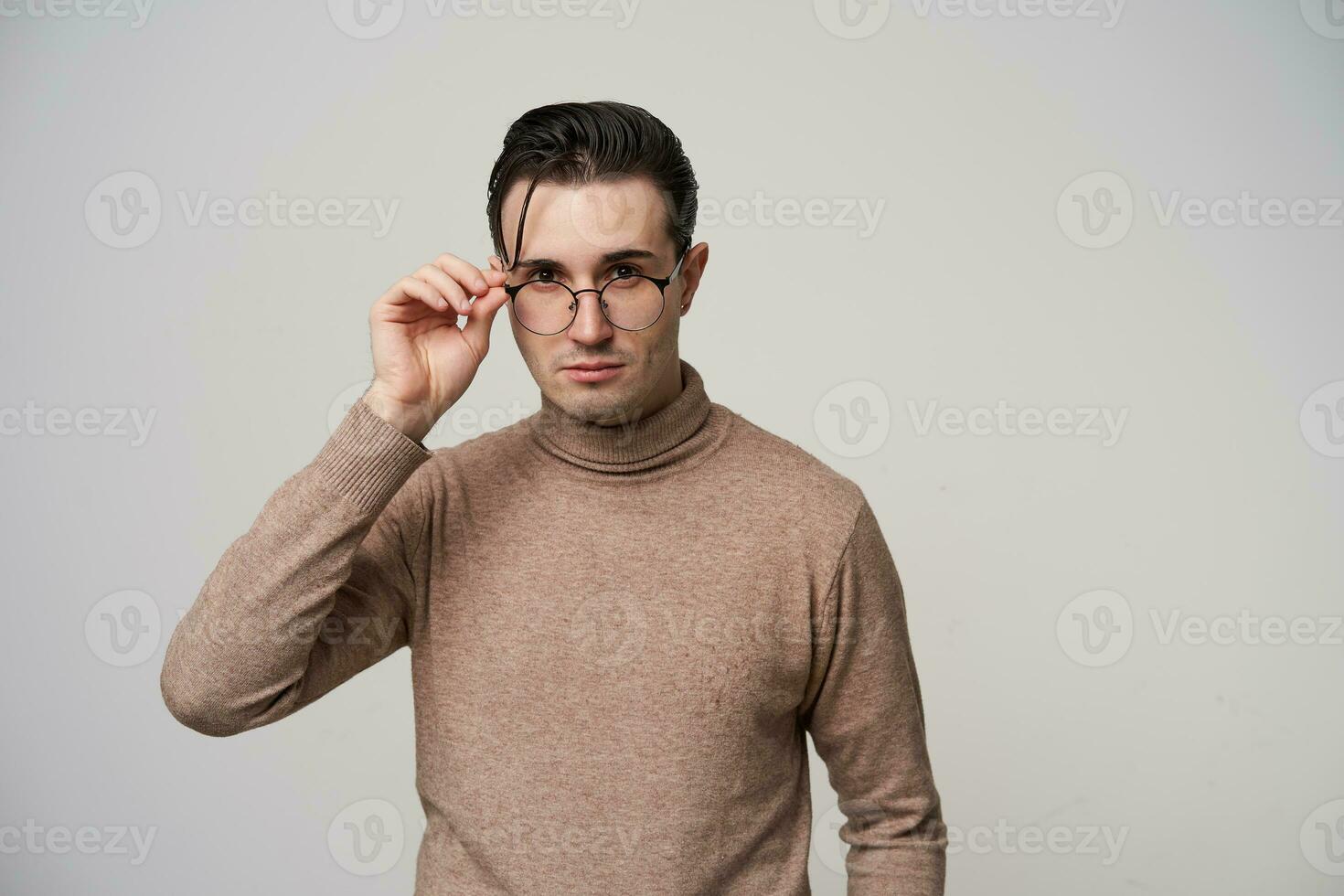 Portrait of pretty young dark haired male hipster in glasses looking to camera with calm face while standing over white background, wearing beige roll-neck sweater photo