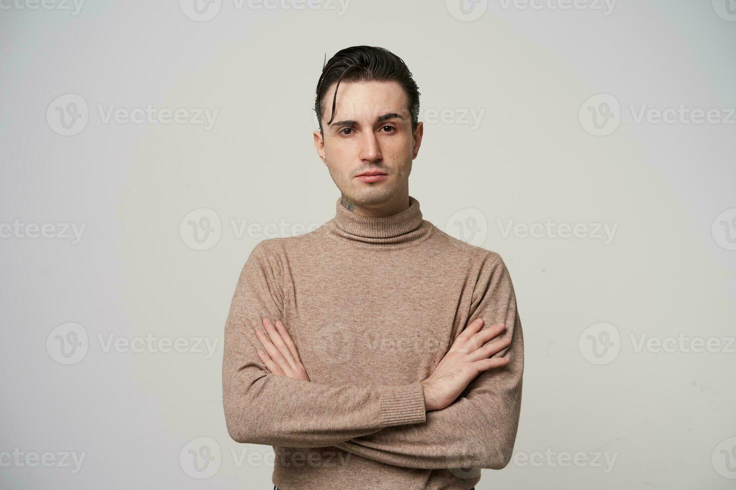 Stylish young pretty dark haired guy with brown eyes keeping hands on his chest and looking severely to camera with raised eyebrow, wearing trendy clothes over white background photo