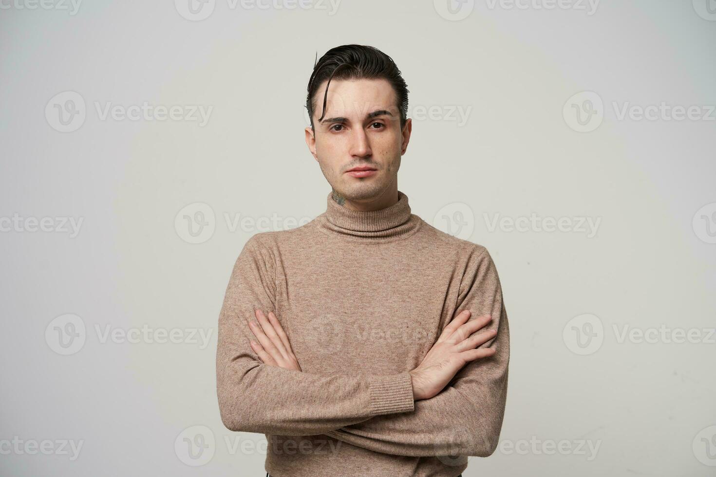 elegante joven bonito oscuro peludo chico con marrón ojos acuerdo manos en su cofre y mirando severamente a cámara con elevado ceja, vistiendo de moda ropa terminado blanco antecedentes foto