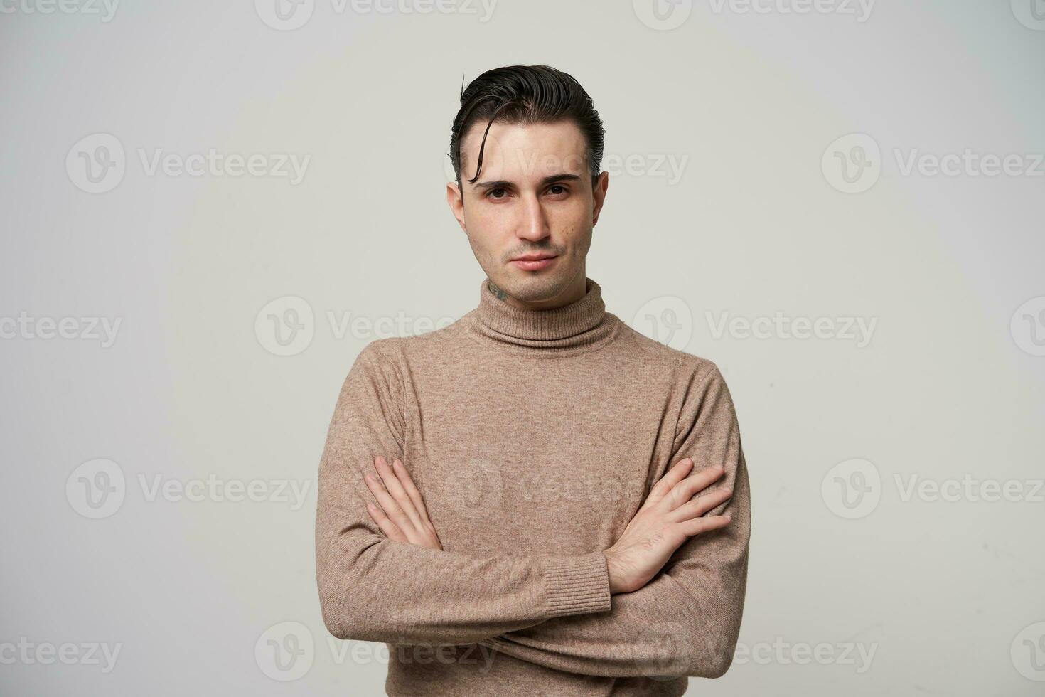 Indoor photo of pretty young brunette male in beige roll-neck sweater crossing hands and looking at camera with folded lips, dressed in trendy clothes while standing over white background