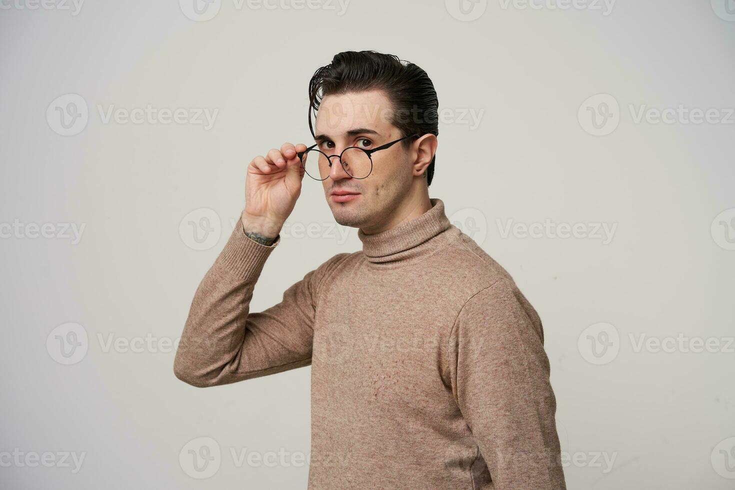 Portrait of handsome brown-eyed young brunette man with trendy haircut holding his eyewear with raised hand and looking at camera with calm face, isolated over white background photo