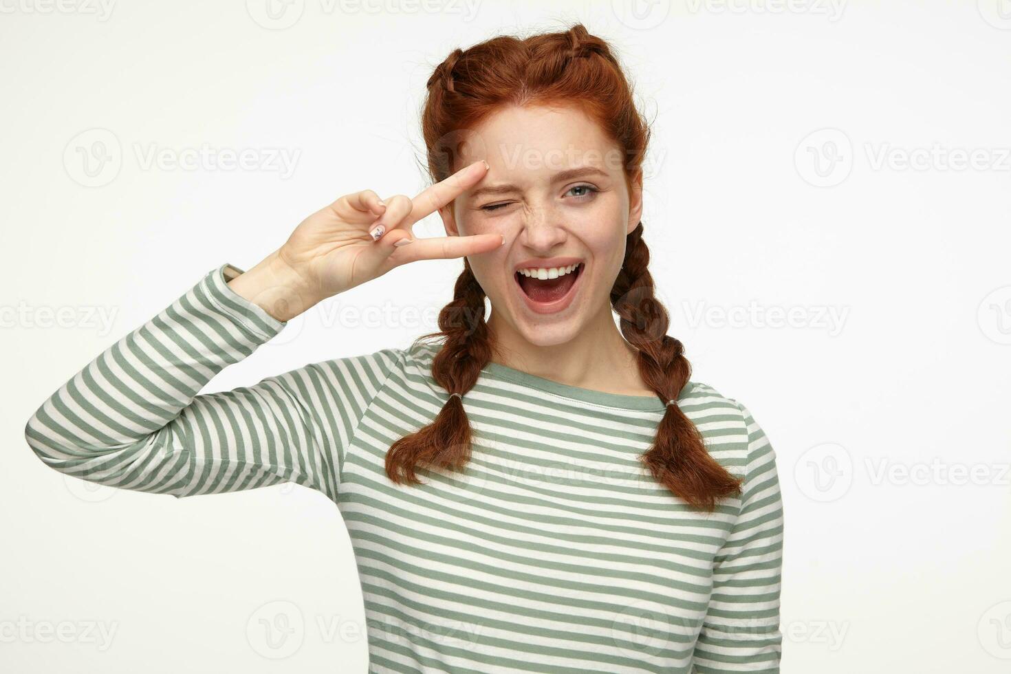 portrait of young ginger female standing over white studio background shows victory sign into camera, wink and smiles broadly photo