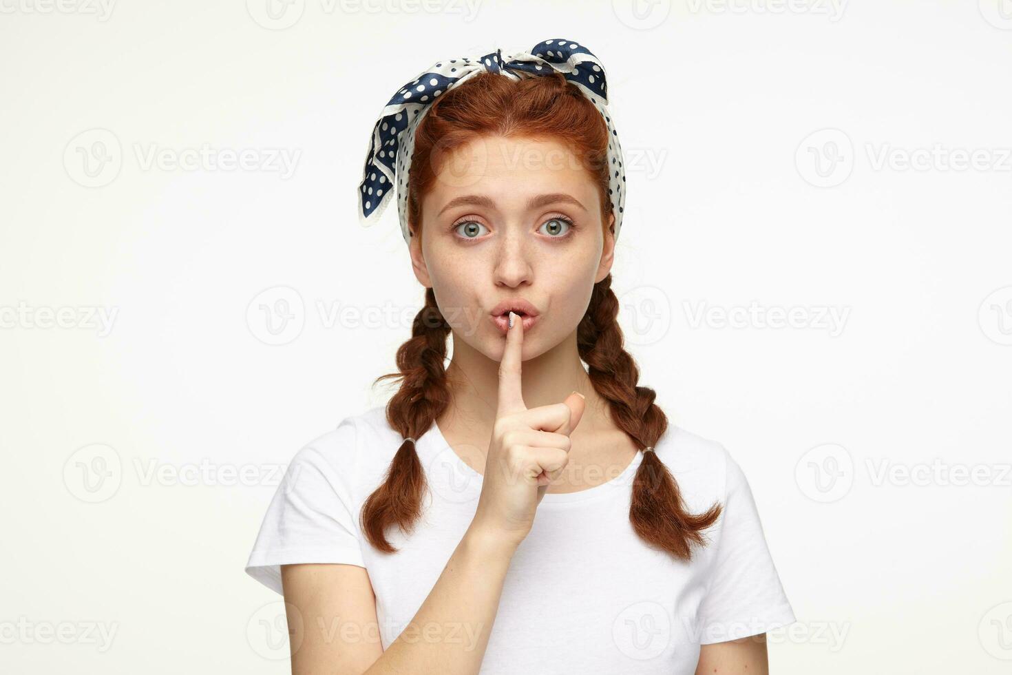 portrait of young ginger female standing over white studio background shows silence gesture with socked facial expression photo