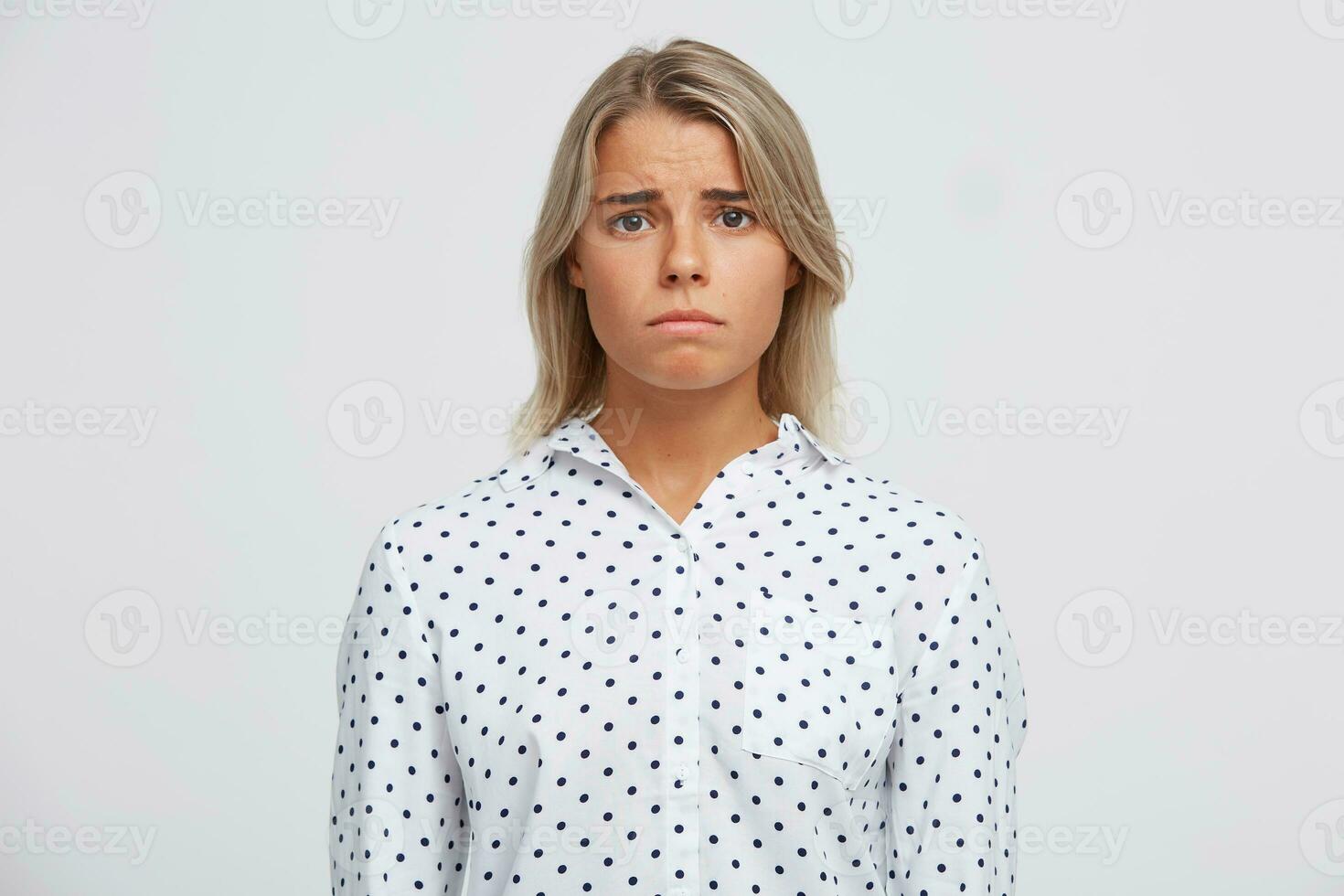 Closeup of sad depressed blonde young woman wears polka dot shirt feels upset and disappointed isolated over white background Looks directly in camera photo