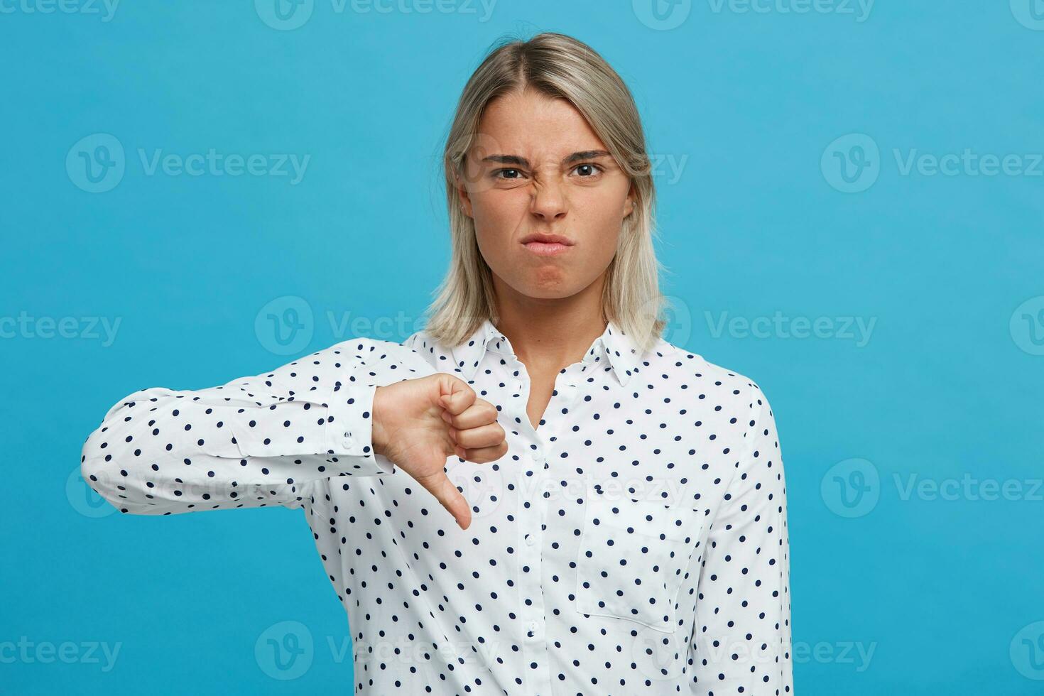 Closeup of unhappy displeased blonde young woman wears polka dot shirt feels dissatisfied and shows thumbs down gesture isolated over blue background photo