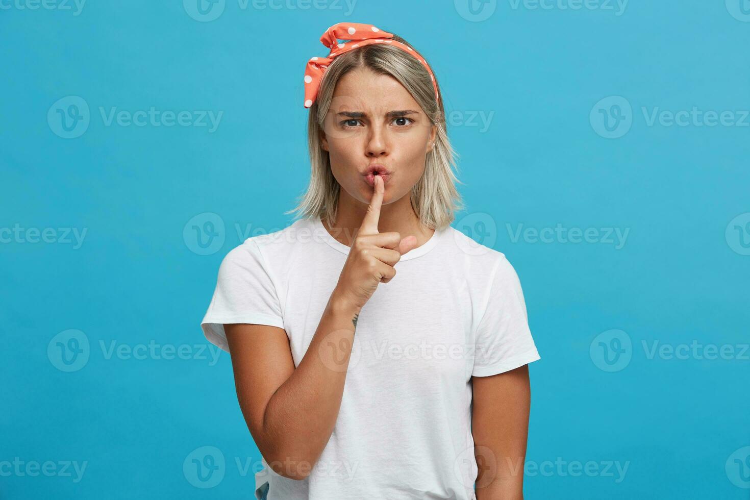 Closeup of serious attractive blonde young woman wears white t shirt and pink headband looks strict and shows silence gesture isolated over blue background photo