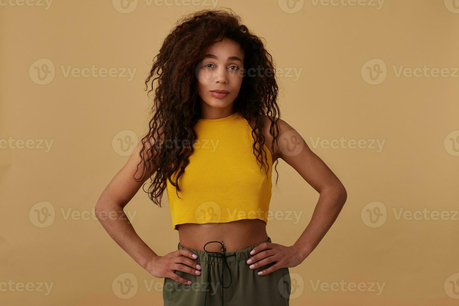 Portrait of young beautiful brunette dark skinned lady with curly brown hair folding hands on waist while posing against beige background in yellow top photo
