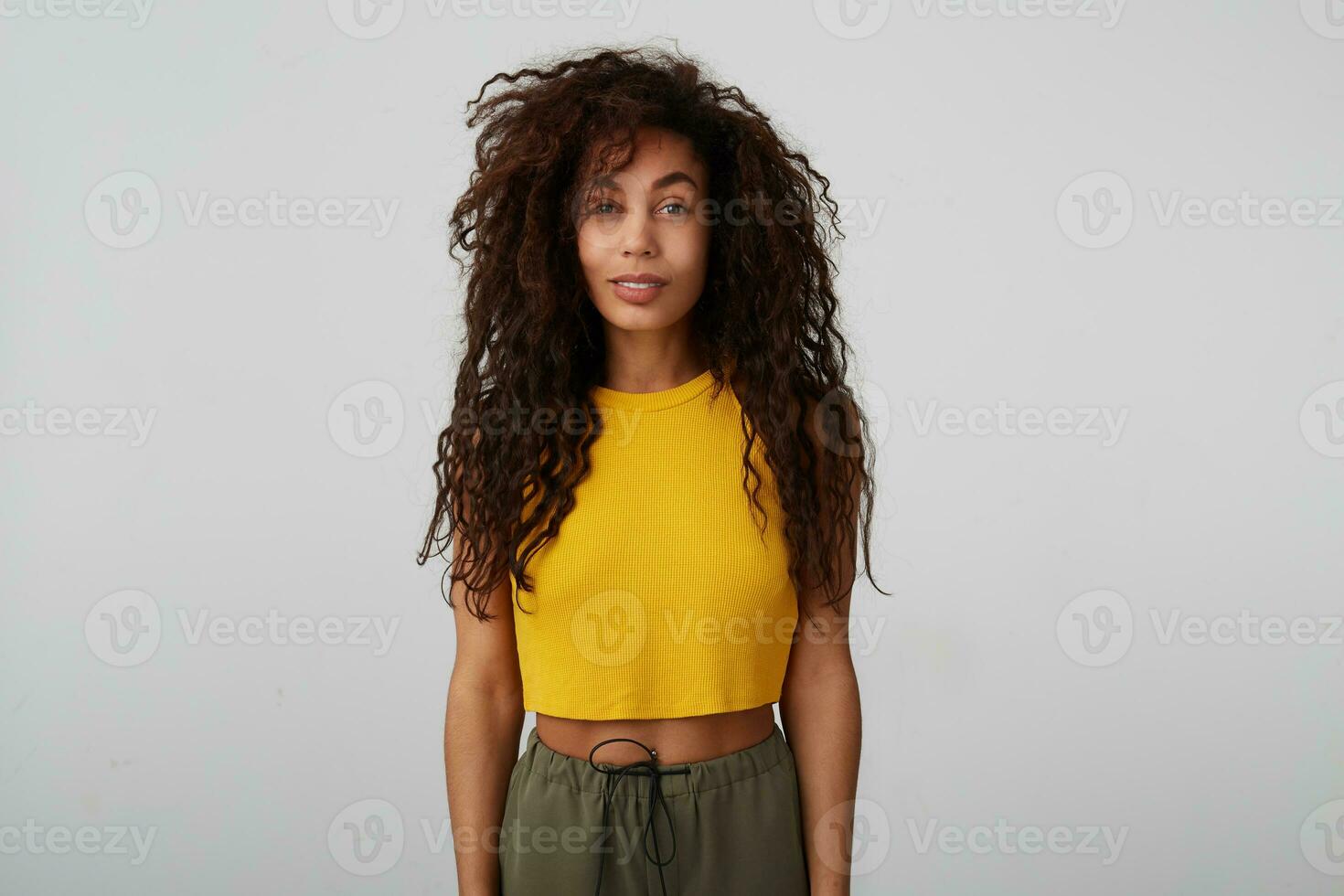 Positive young attractive brunette dark skinned female with wild curly hair keeping hands along body while posing over white background in casual clothes photo