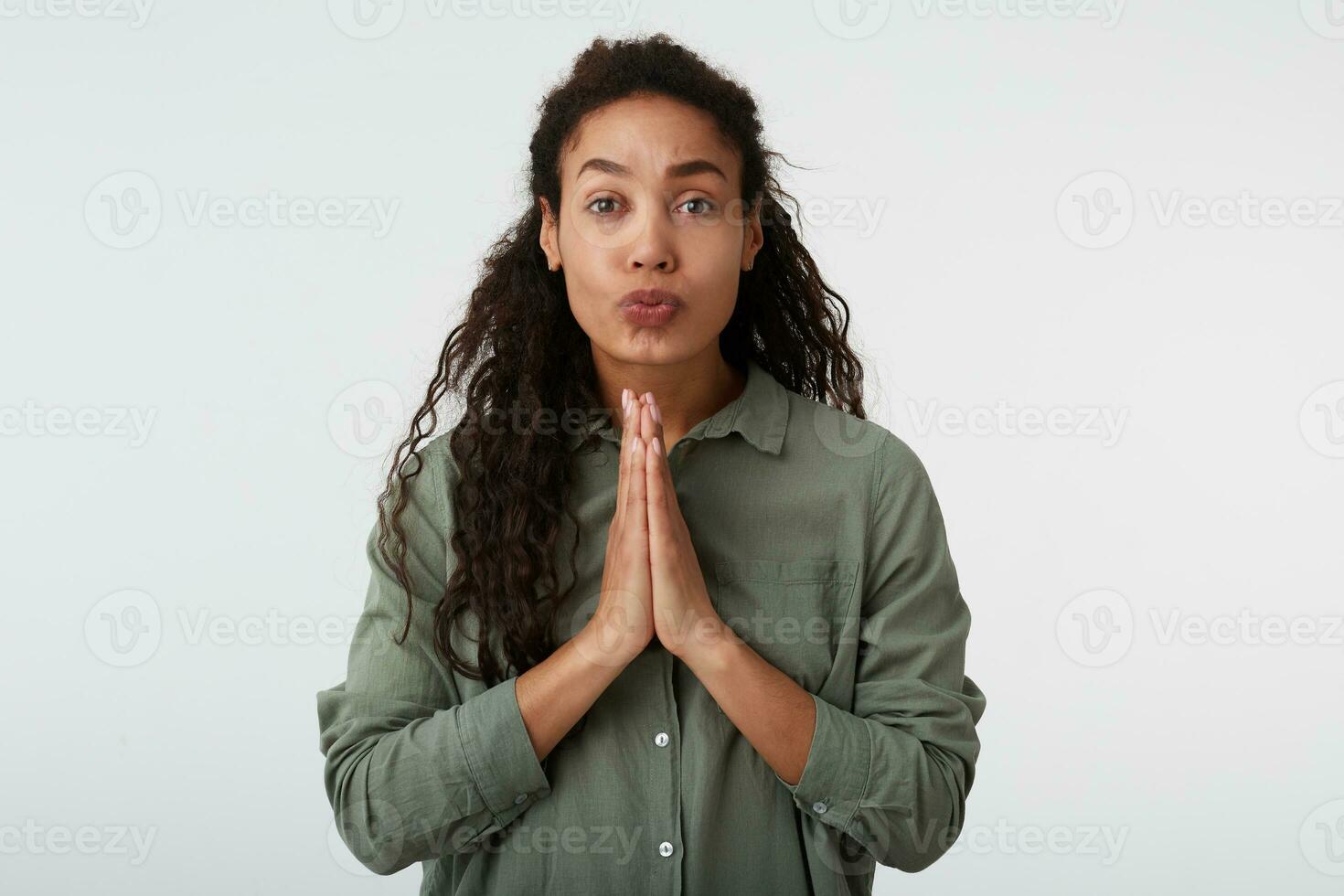 Portrait of hoping brown-eyed curly brunette dark skinned female with casual hairstyle keeping raised palms together and pursing her lips, isolated over white background photo