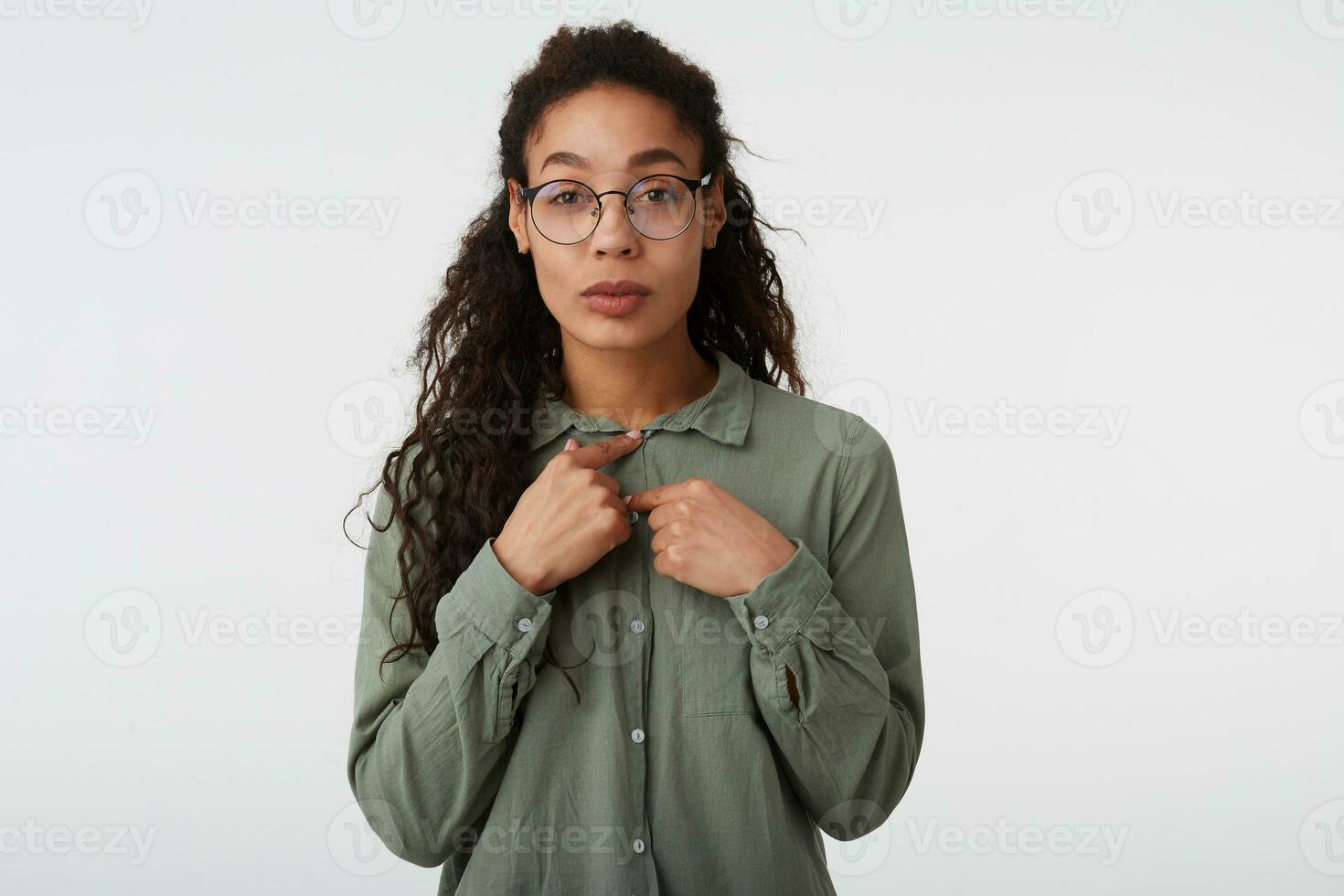 retrato de atractivo joven oscuro peludo Rizado dama con oscuro piel mirando a cámara con doblada labios mientras sujetar botones en su camisa, aislado terminado blanco antecedentes foto