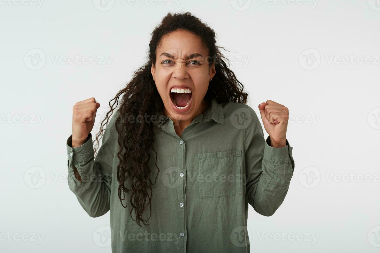 Studio photo of mad long haired curly dark skinned female raising excitedly fists while screaming angrily, dressed in green shirt while posing over white background