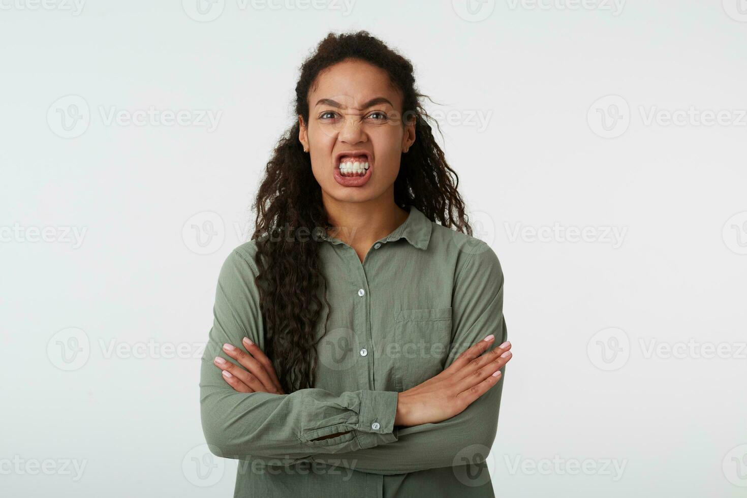 Studio shot of young stressed dark haired curly female with casual hairstyle showing her teeth while grimacing face and keeping hands crossed, isolated over white background photo