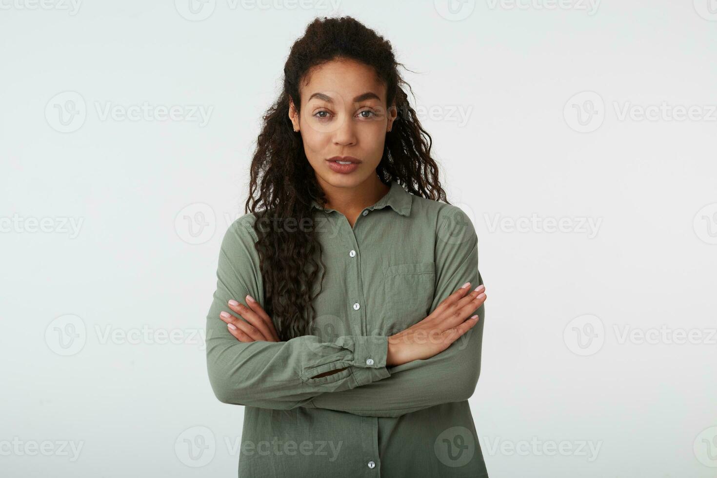 Portrait of young pretty long haired curly dark skinned lady crossing hands on her chest and smiling slightly while posing over white background in casual clothes photo