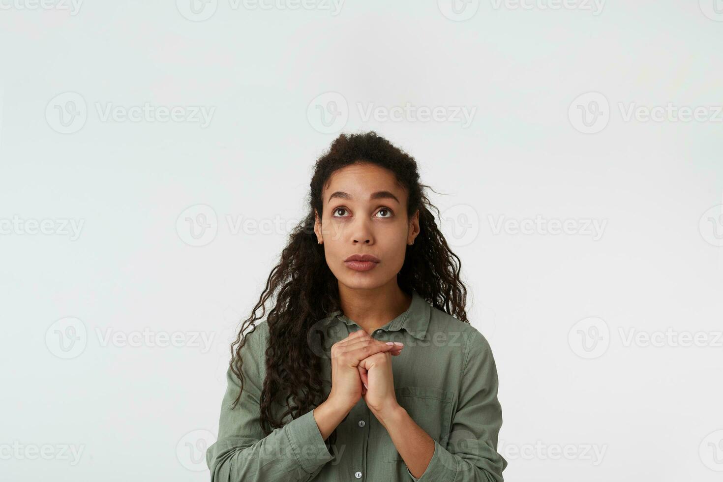 Studio shot of young pretty long haired brunette lady with dark skin folding raised hands while looking hopefully upwards, posing over white background photo