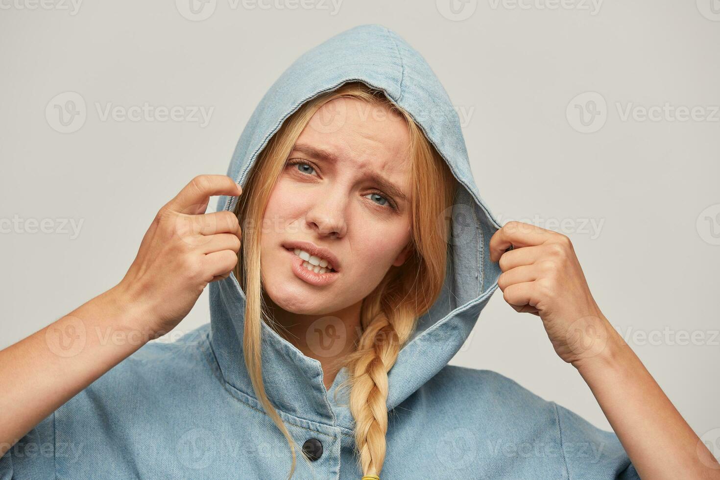 Portrait of beautiful blonde young girl, hands keep hood, looks distressed, wears jeans oversize coat, feeling upset about of bad autumn weather bites her lip isolated over white background photo