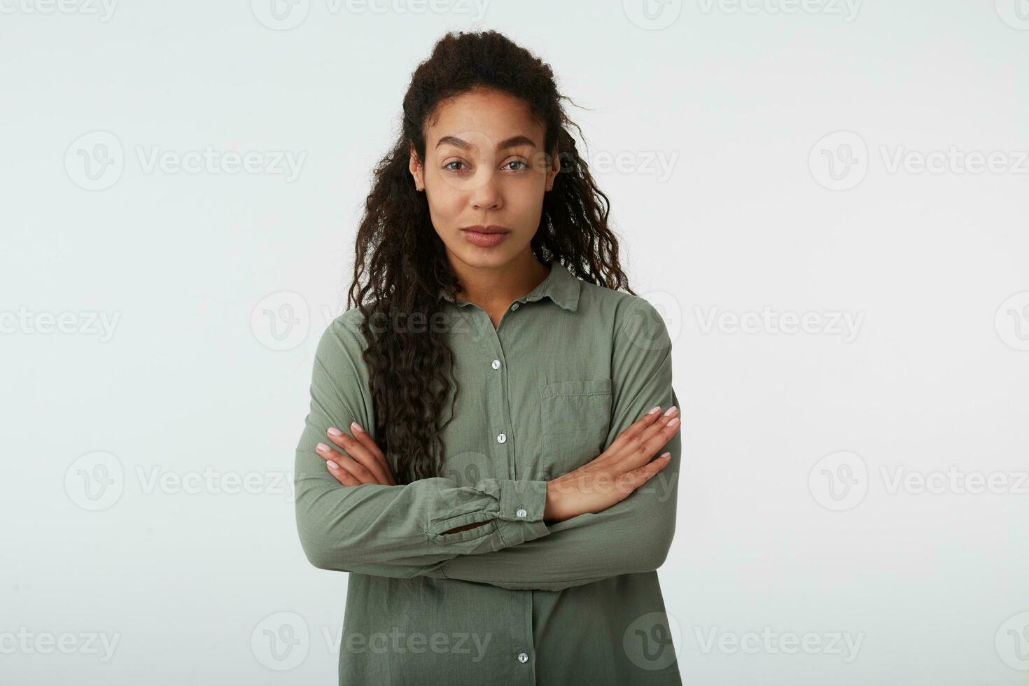 Indoor shot of young attractive brown-eyed curly dark skinned female folding her hands on chest while looking at camera, isolated over white background photo
