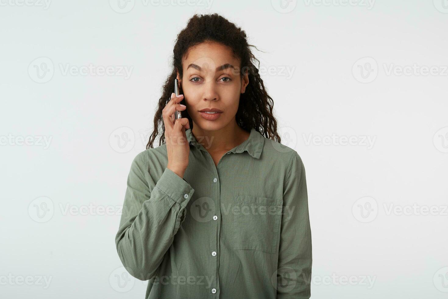 Charming young brown-eyed curly brunette female with dark skin looking calmly at camera while making call with mobile phone, isolated over white background photo
